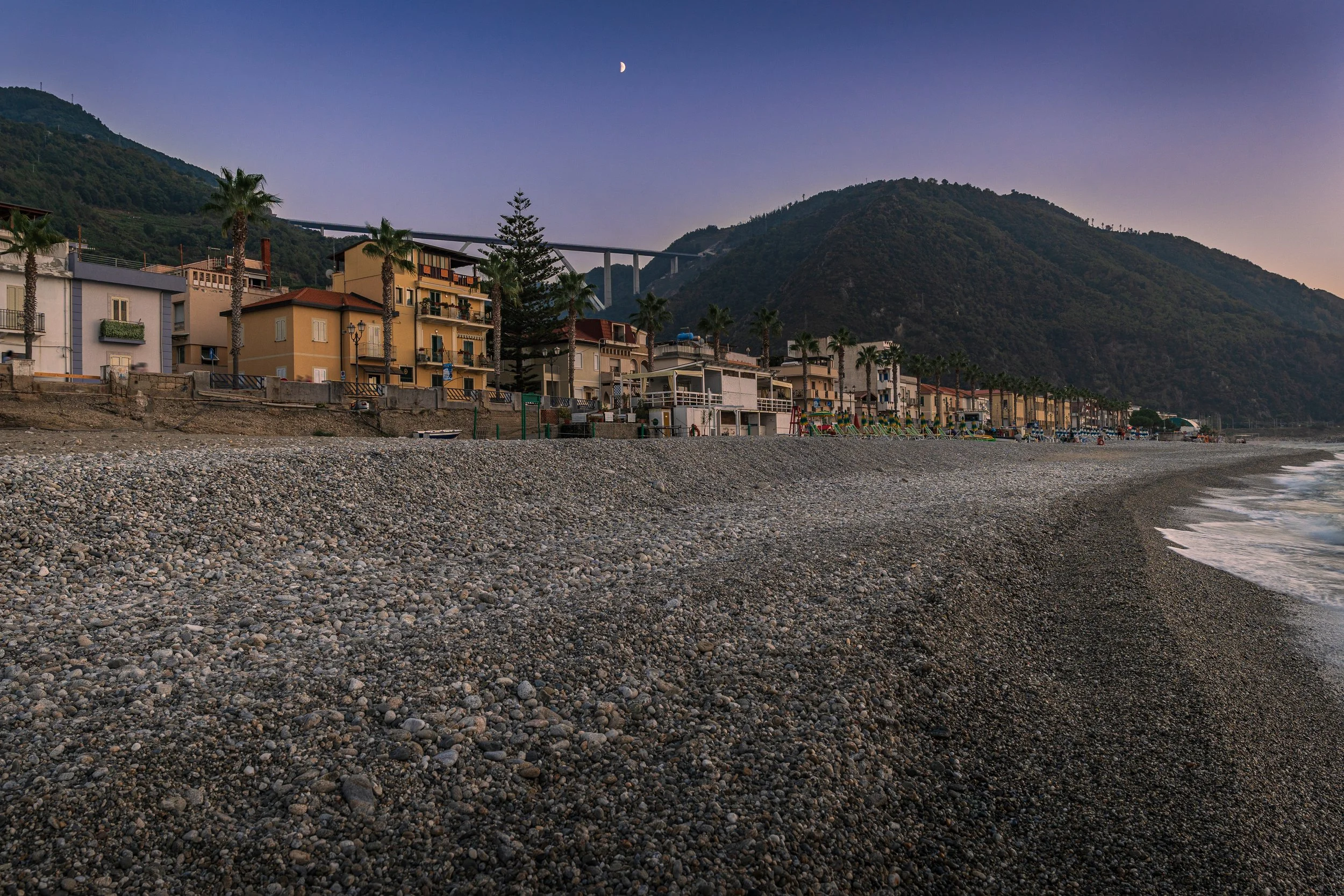 The rocky beach at Bagnara Calabra, Italy, looking toward the Lungomare, lush green mountains, and the A2 Ponte Sfalassà di Bagnara.