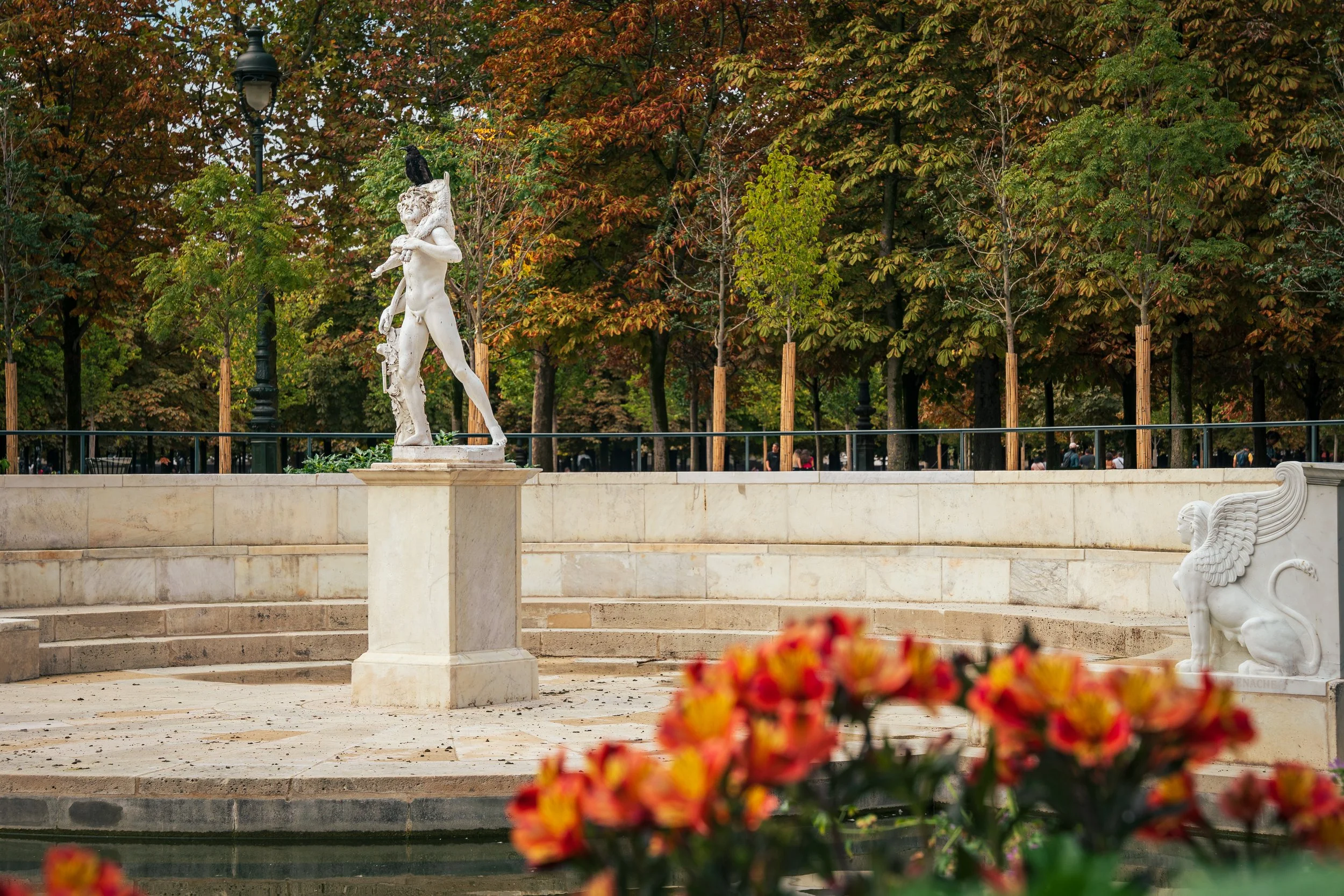 A statue in the Jardin des Tuileries (Tuileries Garden), Paris, France. The leaves are beginning to turn for the fall, though some trees have green leaves. The statue is surrounded by small, amphitheater-like seats, and red and yellow flowers are out