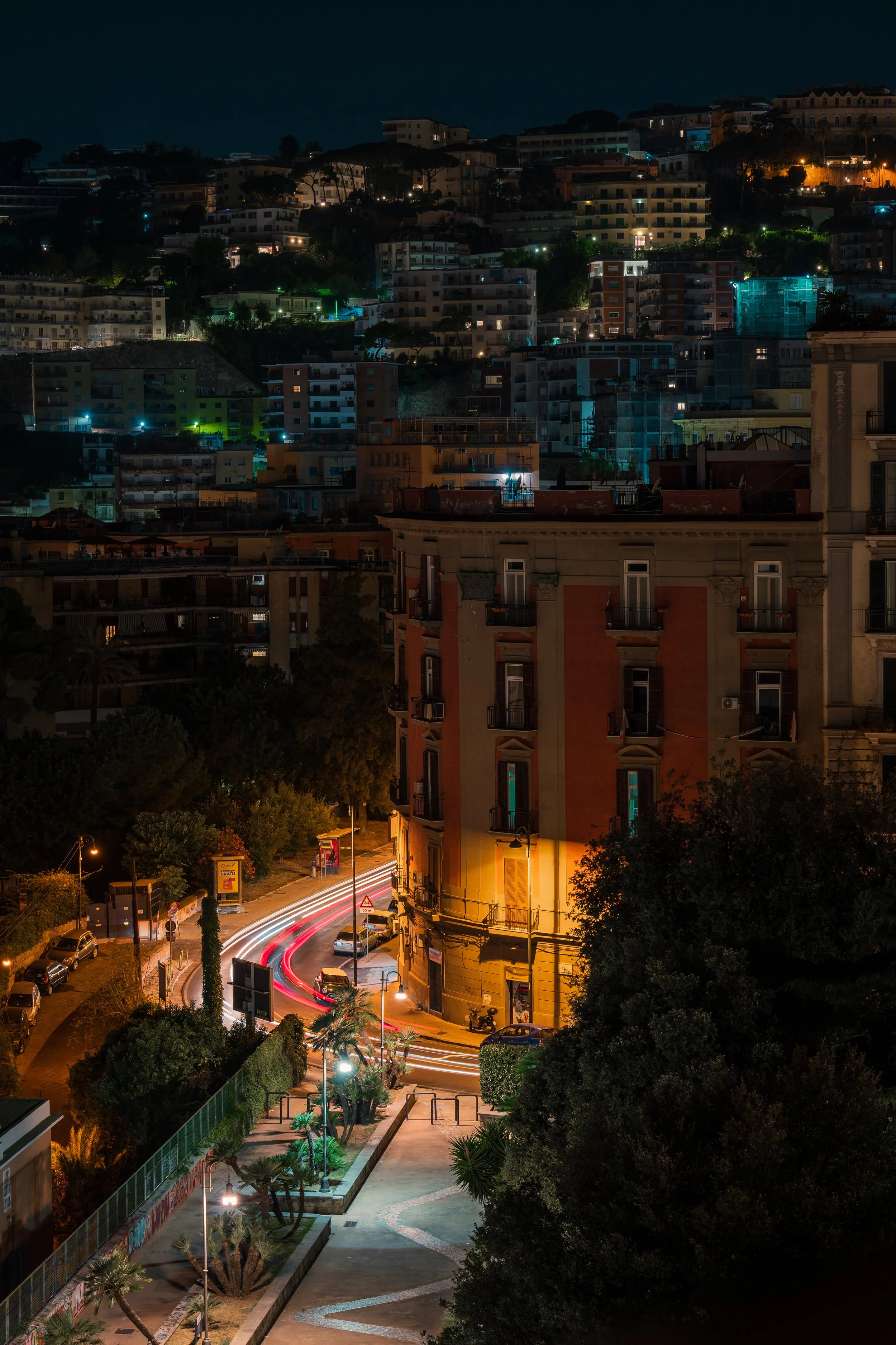 Naples, at night, from the balcony in our Chiaia bed & breakfast. The buildings are alight with orange and blue lights, and we can see car trails in the foreground.