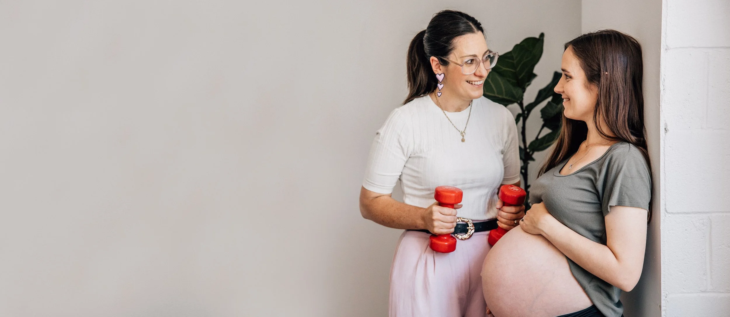 A pregnant woman with long brown hair leaning against a wall smiling at a woman with glasses and dark hair, holding red dumbbells, in an indoor setting.
