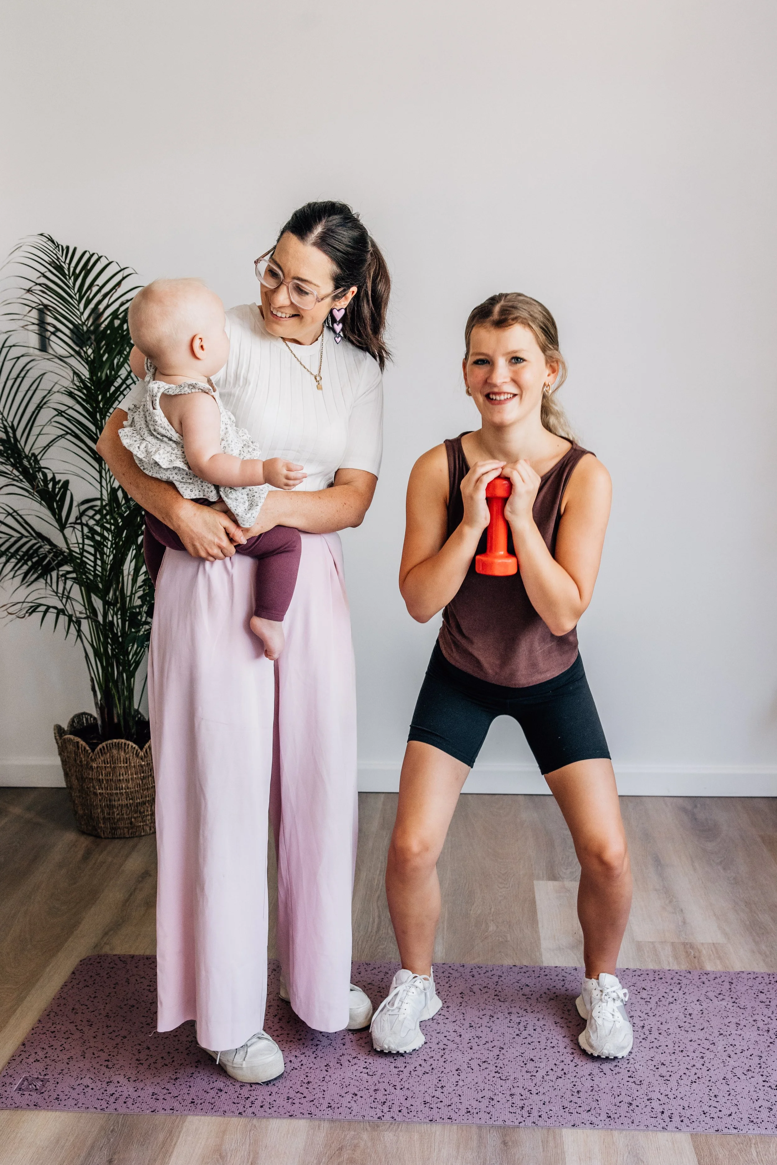 A woman holding a baby and a young woman holding a dumbbell during a fitness session indoors.