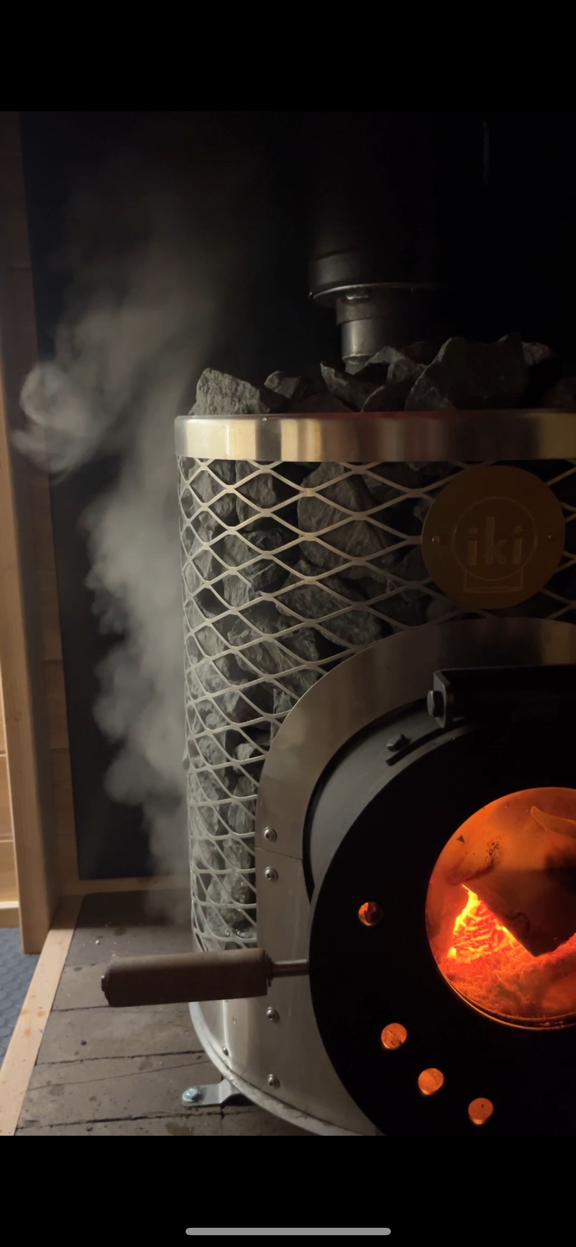 A wood-burning stove with glowing embers and flames inside, surrounded by a metal chimney and a container of rocks, with smoke coming out.