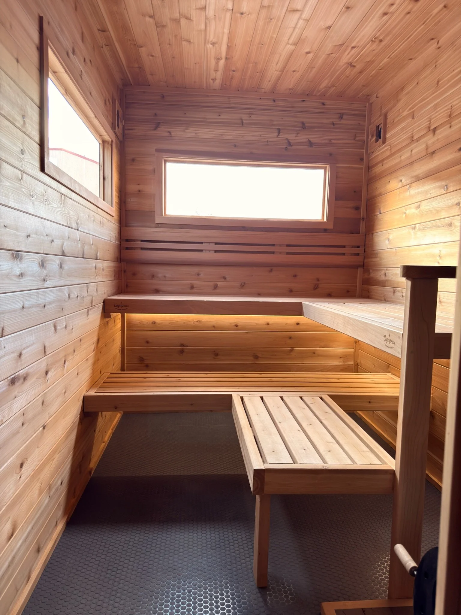 Inside a small wooden sauna with natural wood paneling, a wooden bench, and two windows providing natural light.