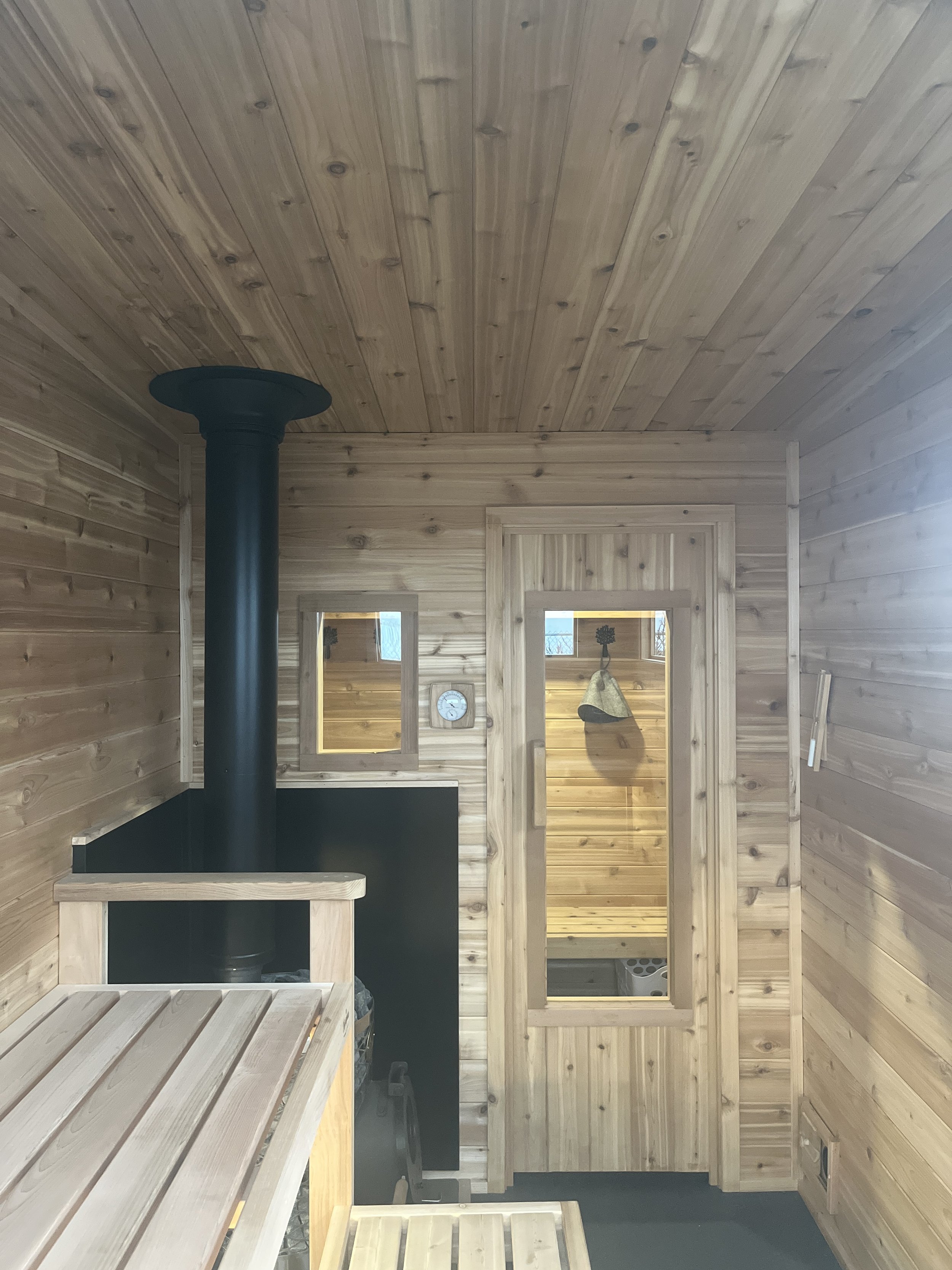 Interior of a small wooden sauna room with wood-paneled walls and ceiling, a black stove pipe, and a wooden bench. There is a small window and a glass door leading to a shower area.