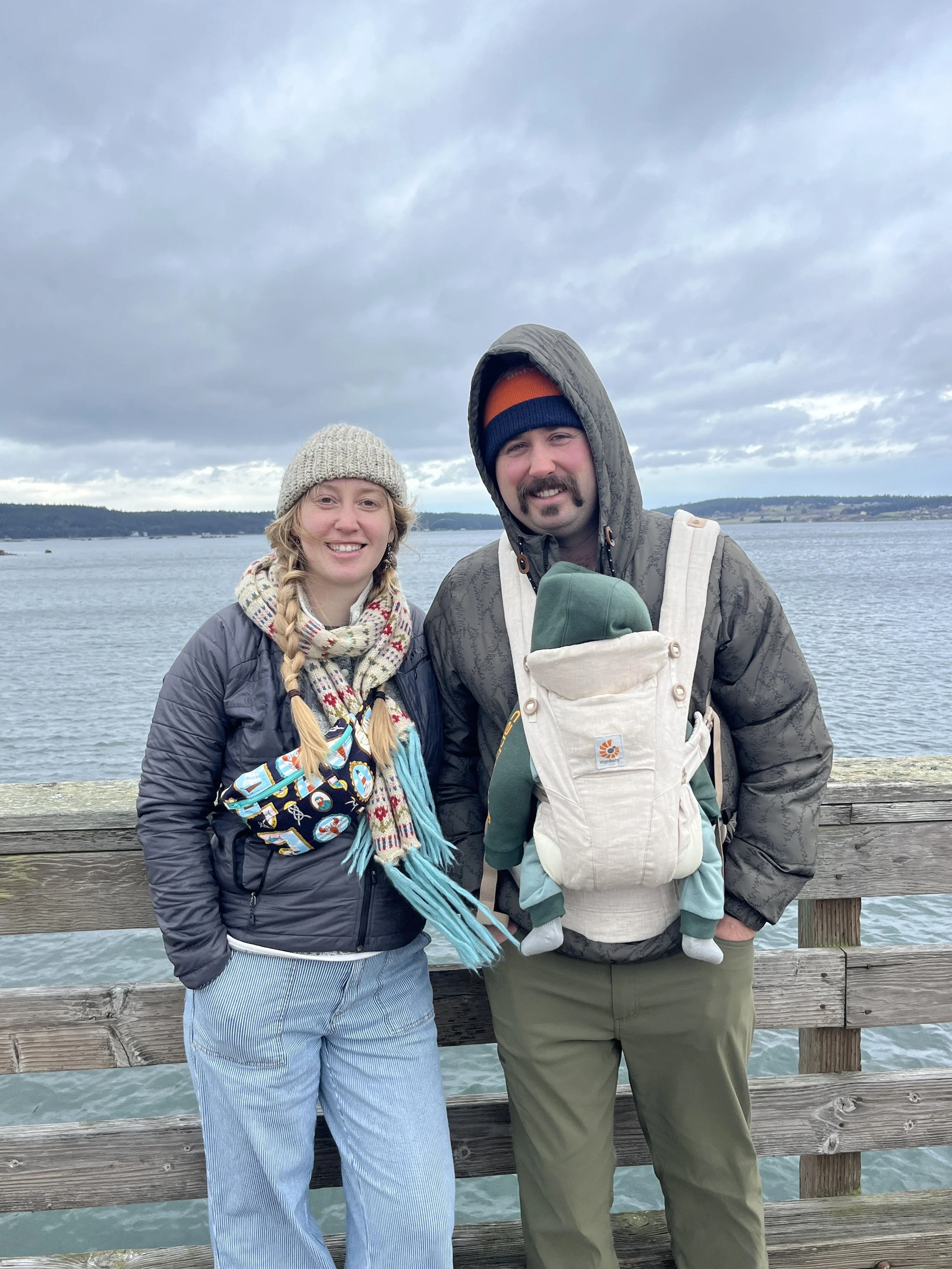 A smiling couple standing on a wooden pier by a body of water, wearing warm outdoor clothing and a baby carrier with a child bundled inside.