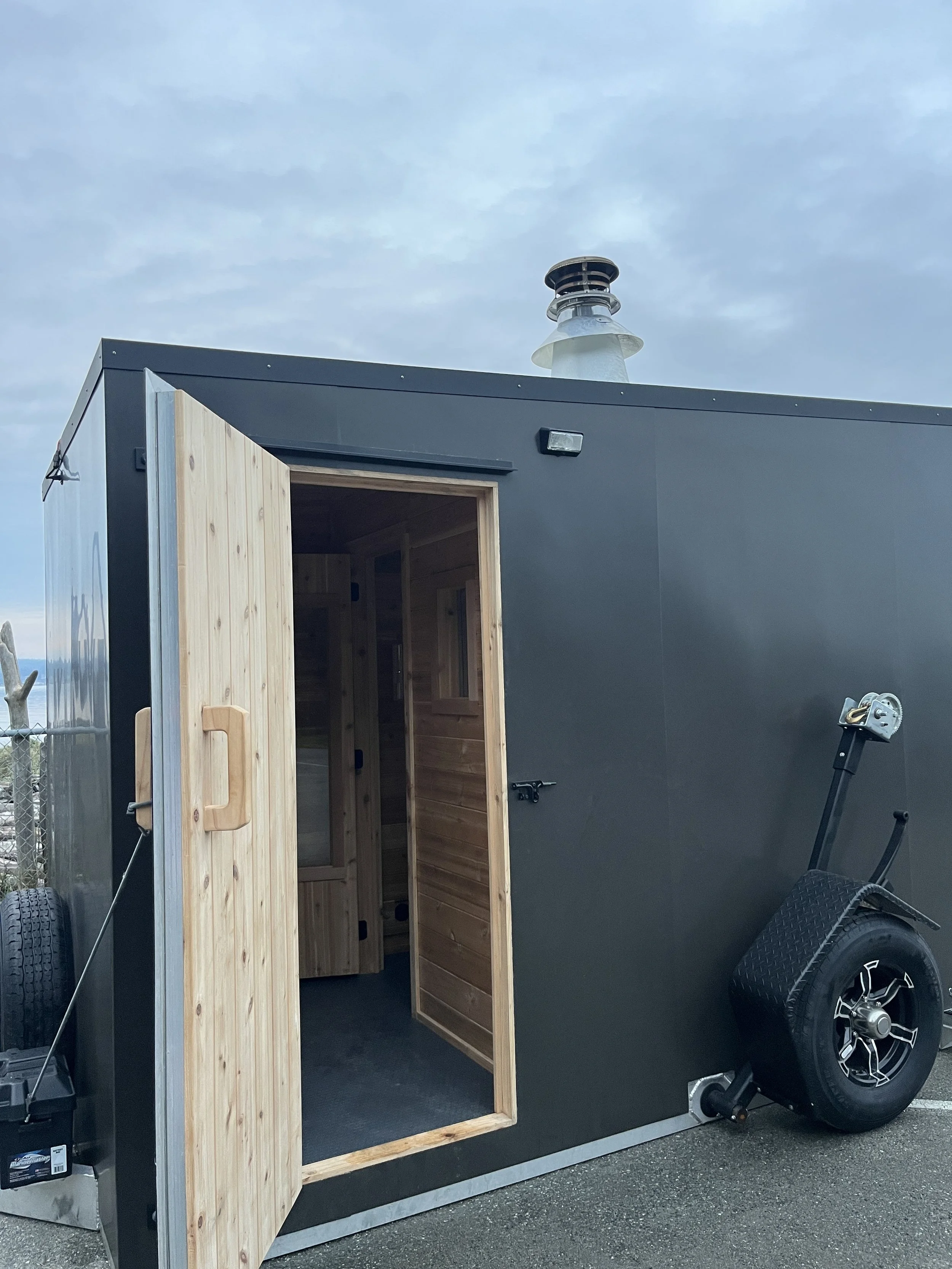 A black tiny house trailer with an open wooden door, showing a interior with wooden walls, a small window, and a gray floor. There is a spare tire attached to the trailer and a wheelbarrow resting against it, with a cloudy sky overhead.