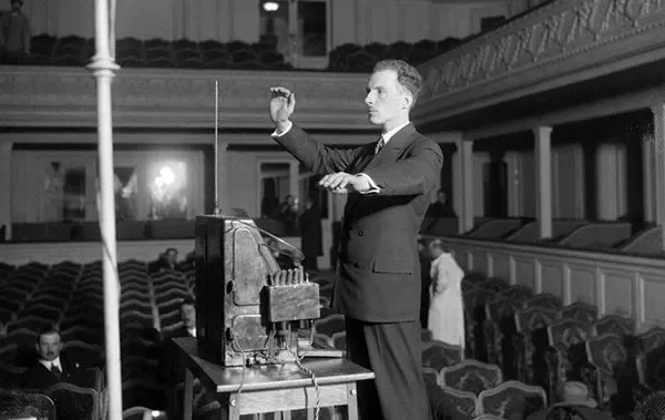 A conductor standing in front of an orchestra conducting with a baton in a large auditorium or concert hall.