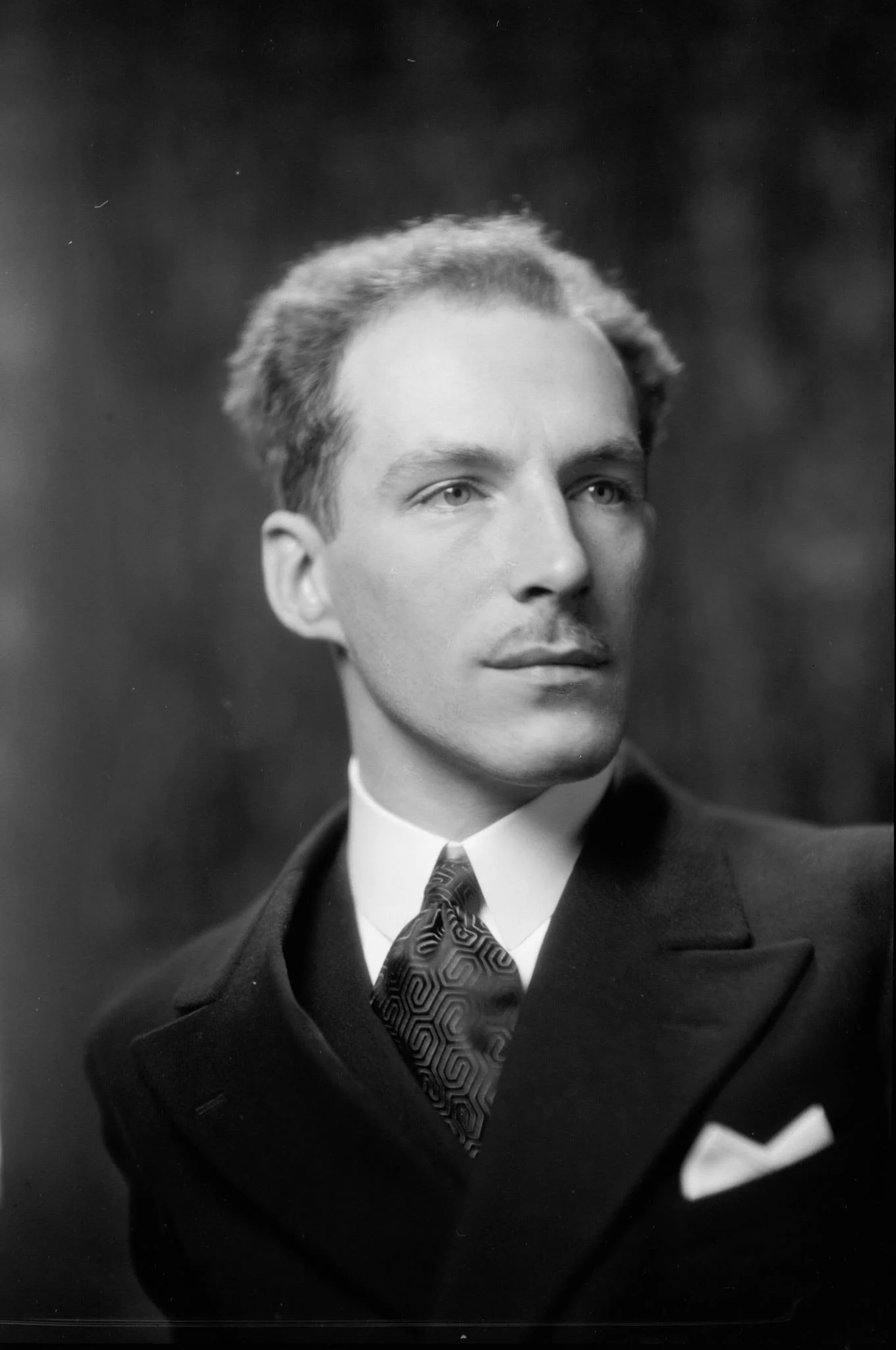 Black and white portrait of a man in a suit with a patterned tie and pocket square, looking to the side with a serious expression.