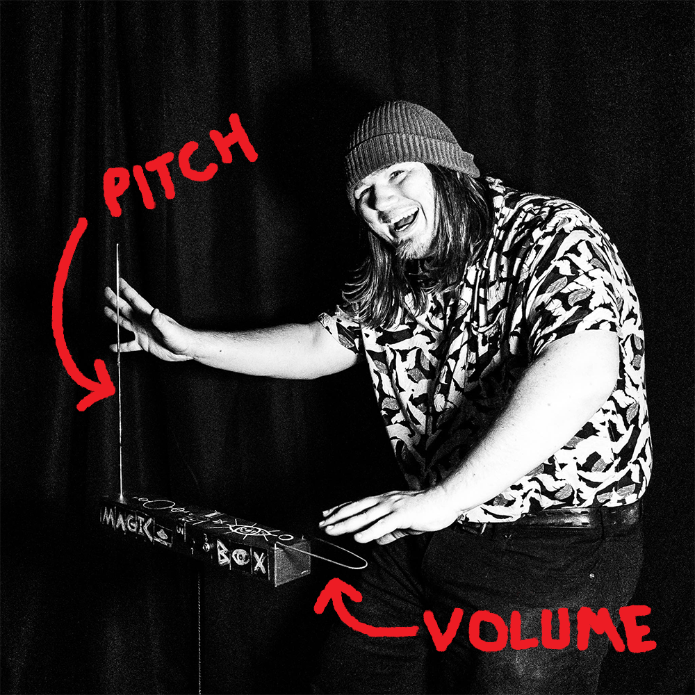 A man with long hair and a beanie, smiling and touching a musical instrument with the words 'PITCH' and 'VOLUME' written in red and arrows pointing to different parts of the instrument.