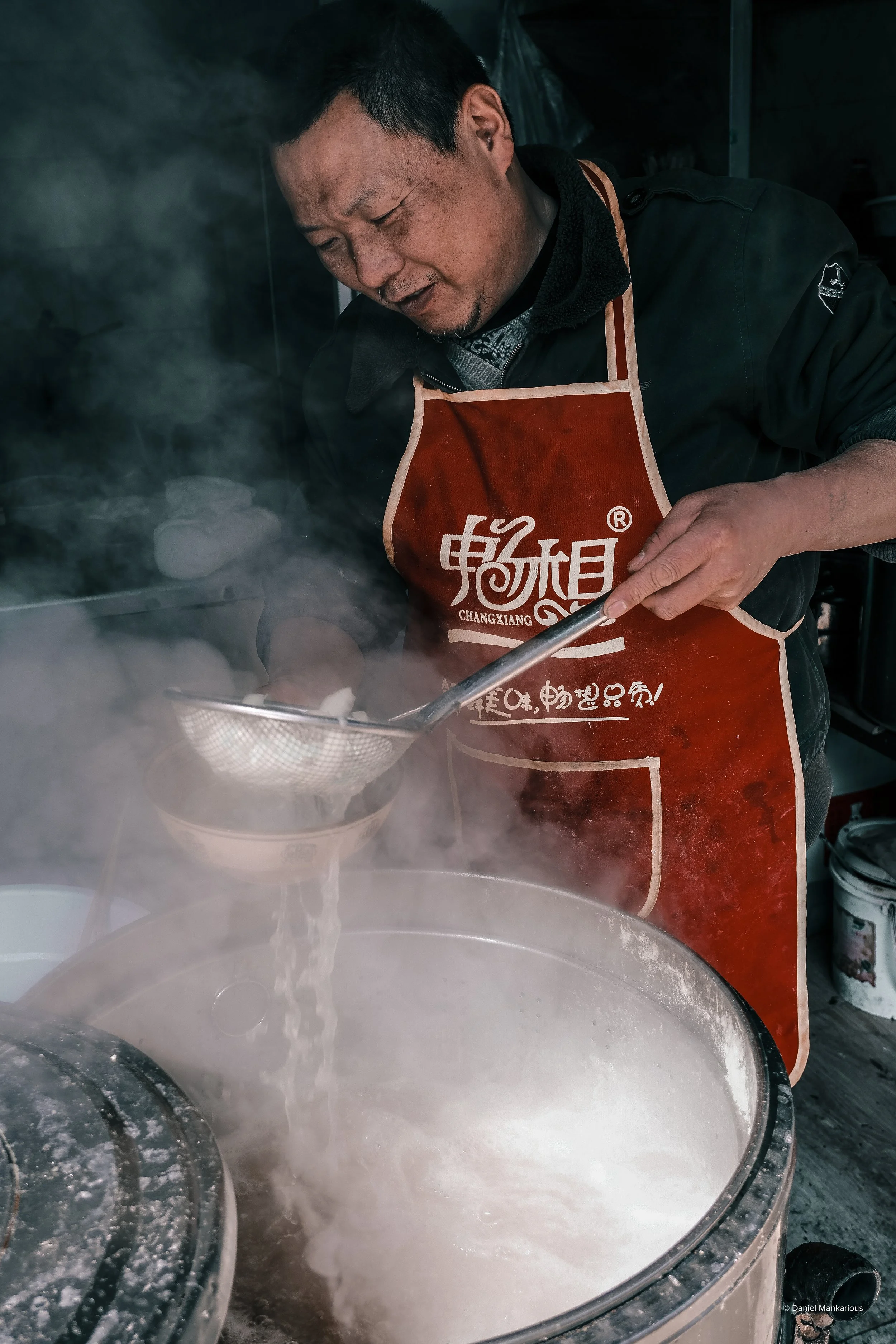 A man wearing a red apron cooking over a large steaming pot, using a skimmer to stir or scoop food.