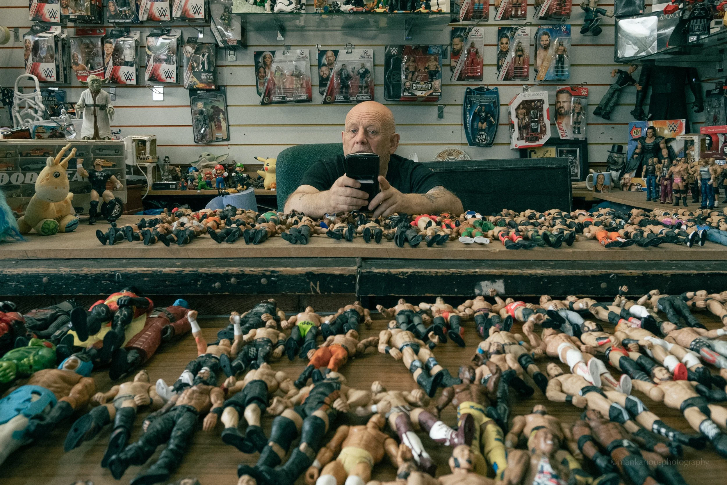 A man with a bald head and tattoos on his arms sitting behind a table filled with numerous toy action figures and dolls in a toy store. The store shelves in the background display more action figures and collectibles.