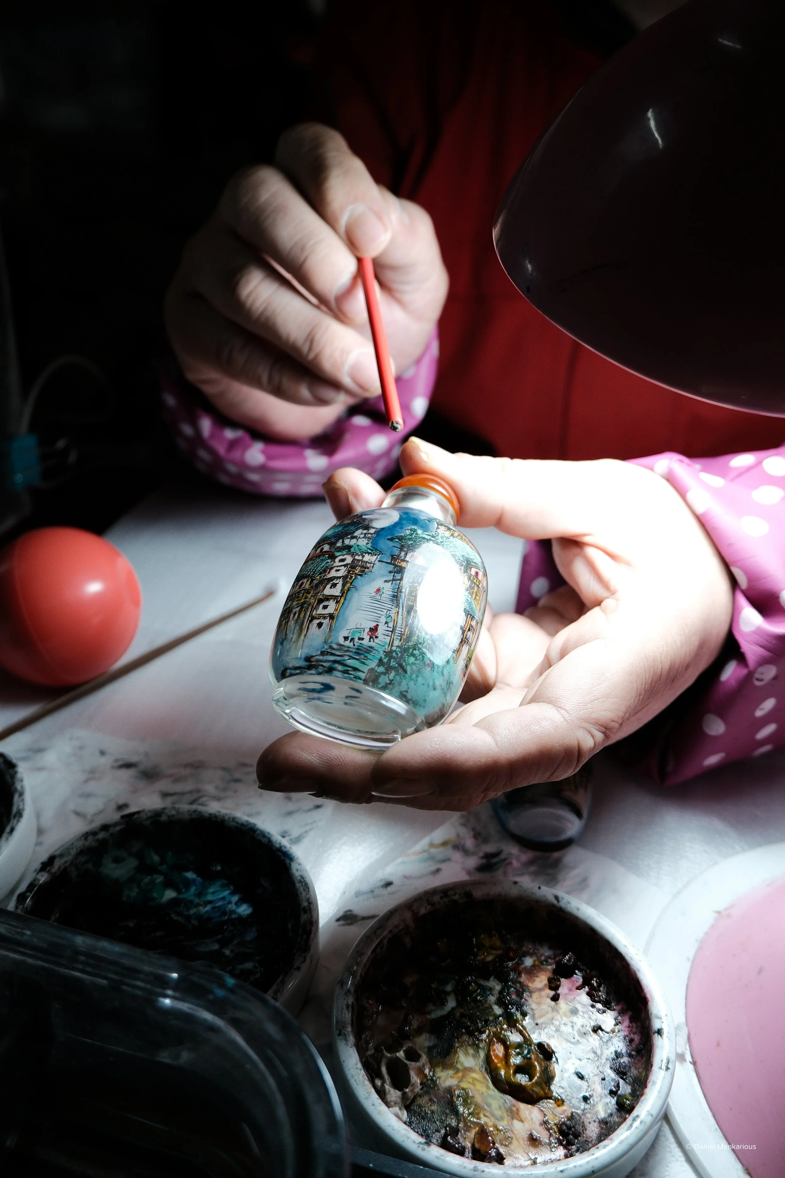 Person painting a detailed scene on a small glass object using a fine brush and red paint, with painting supplies on a work surface.