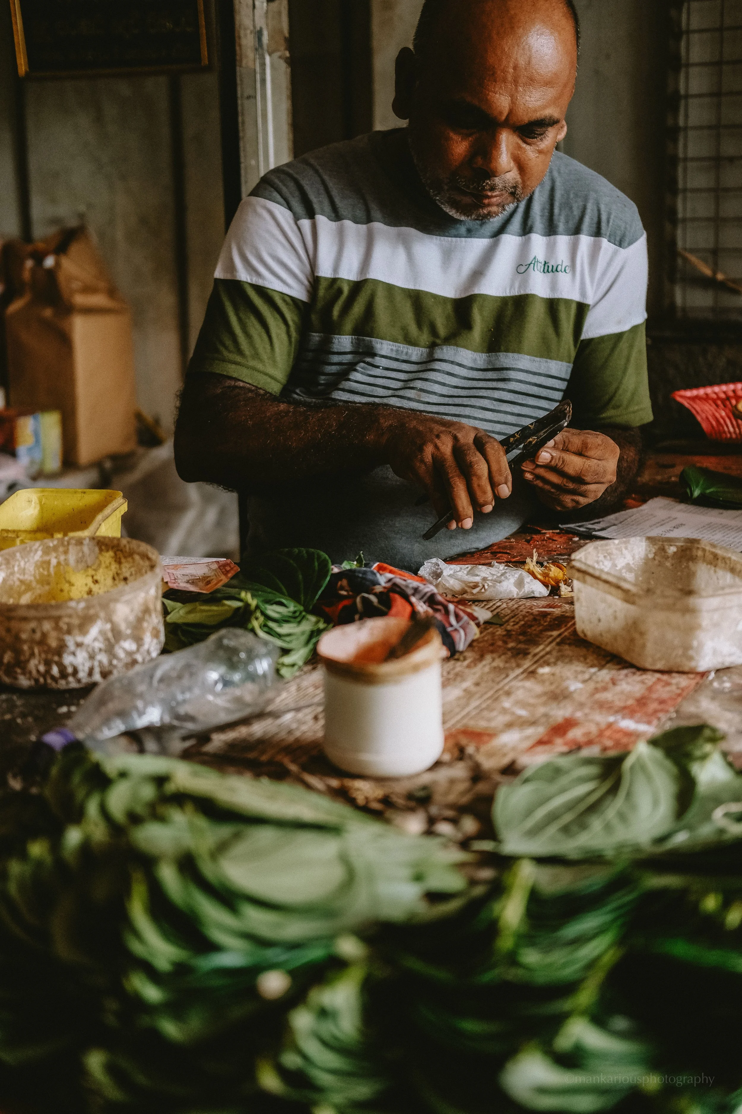 A man wearing a striped T-shirt standing at a wooden table, taking a photo with his smartphone. The table has various vegetable ingredients, and the foreground shows a pile of green leafy vegetables.