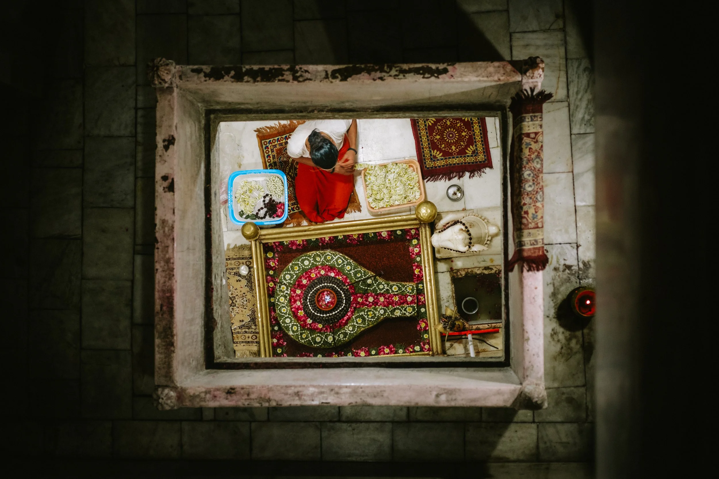 A person with black hair wearing a white top and red skirt is sitting on a rug, preparing flower offerings on trays with white and green flowers, inside a Hindu temple. The photo is taken from a high, top-down perspective through a square opening.