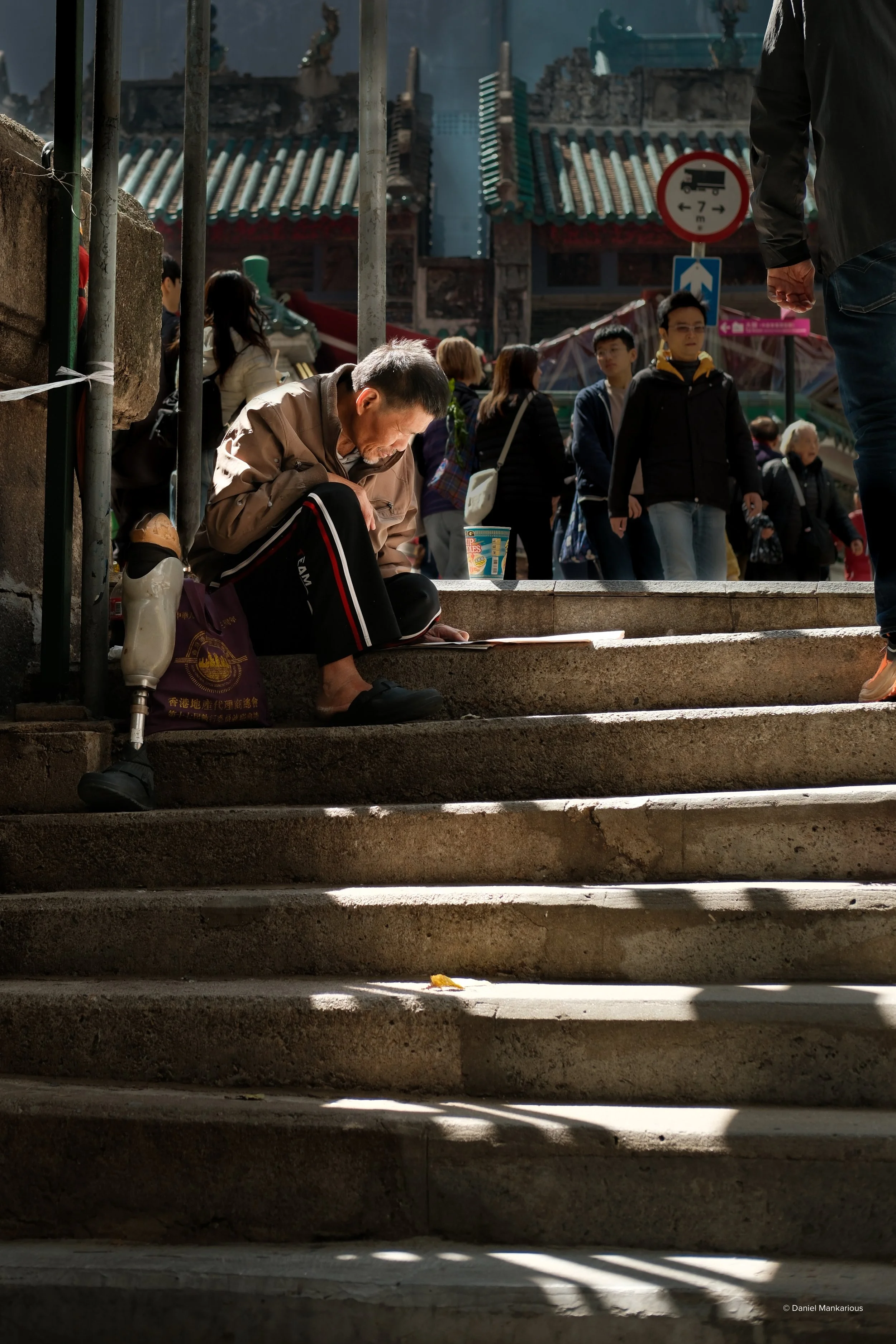 A man with a prosthetic leg, sitting on a staircase, reading or writing on a piece of paper. He is surrounded by a crowd of people in a busy urban setting with traditional Asian architecture in the background.