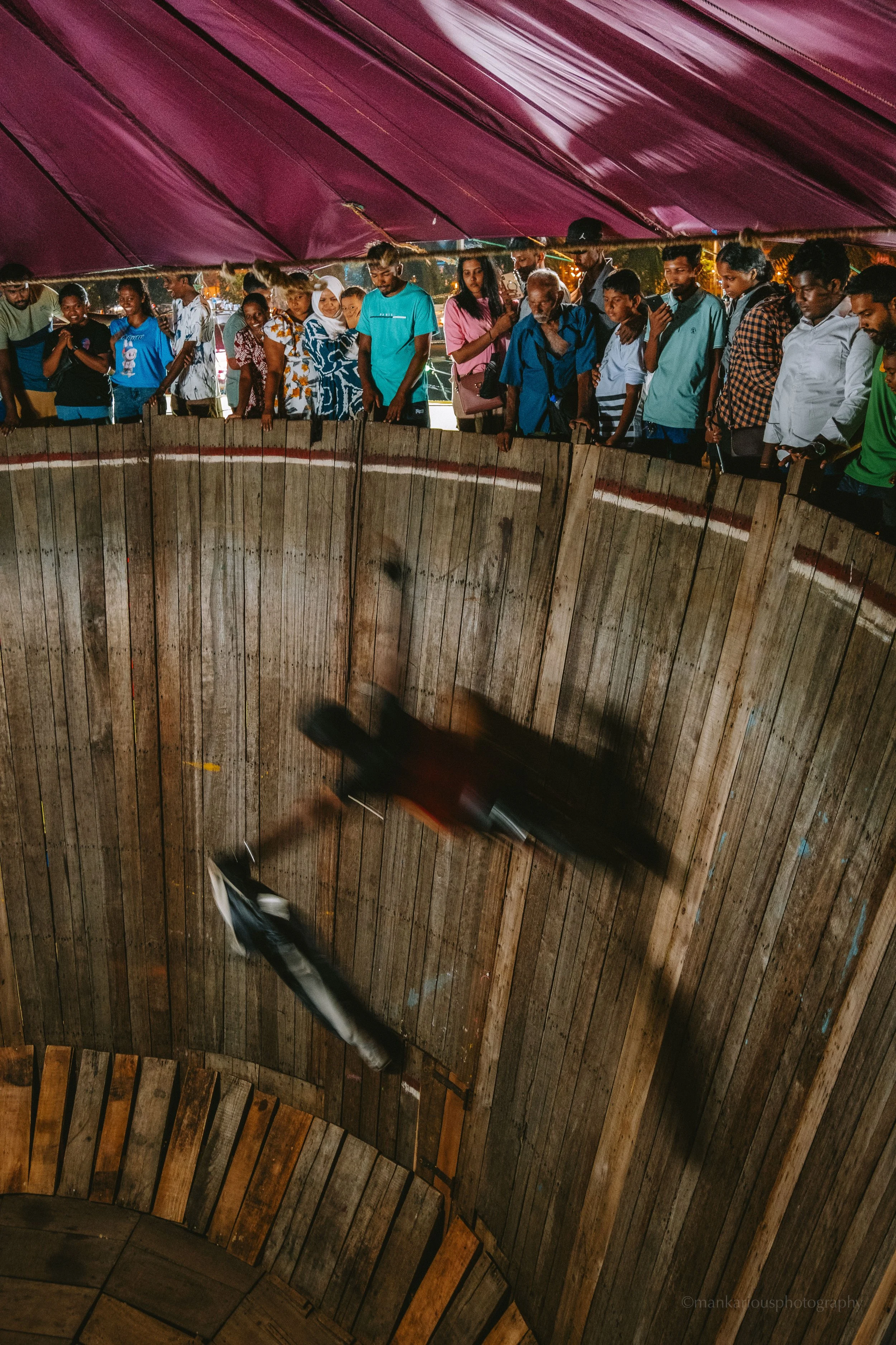 A group of people standing around a wooden wall, watching two motorcyclists perform stunts inside a wooden ramp at a circus or event.