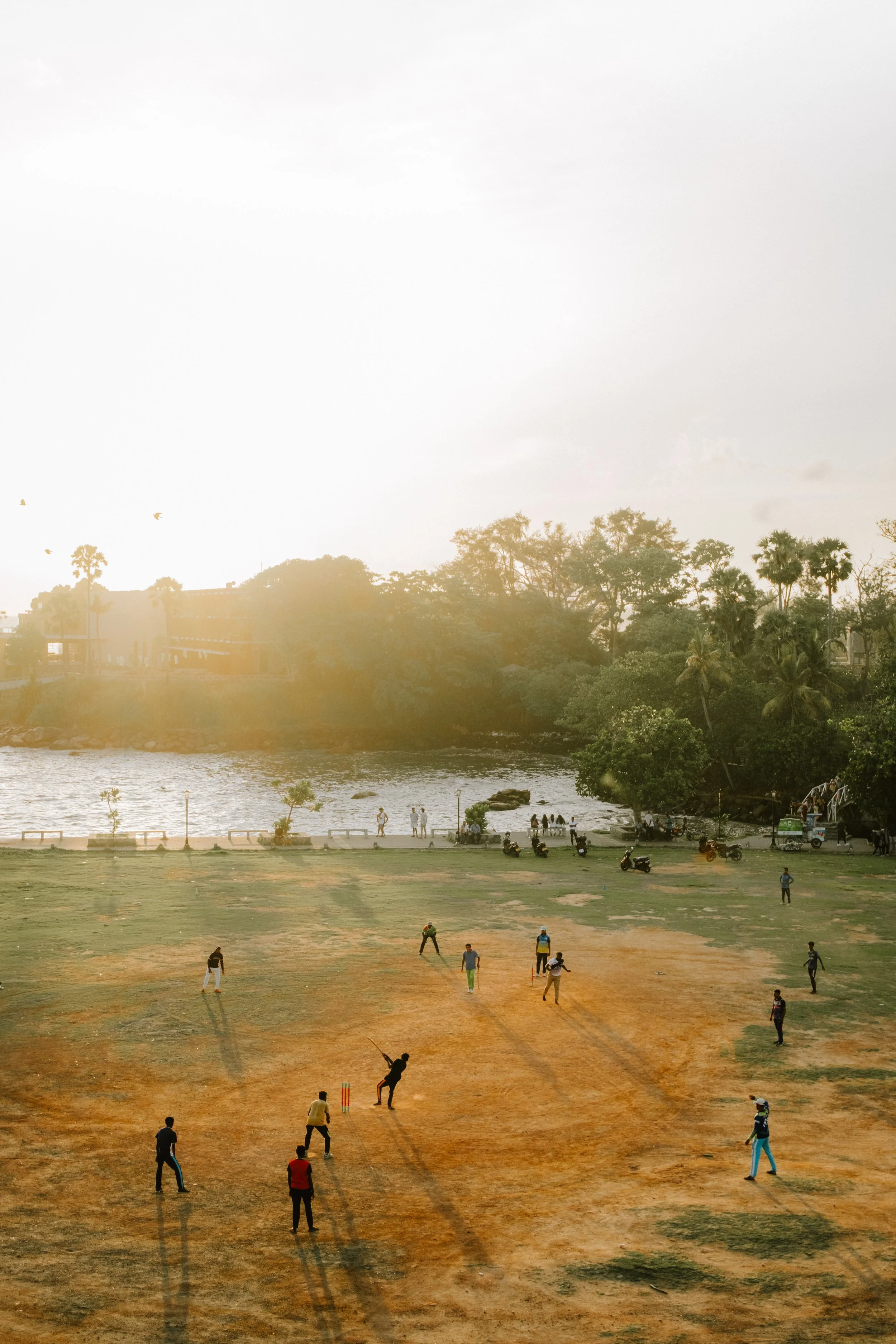 People playing cricket on a sandy field near a river, with trees and buildings in the background, under a bright, cloudy sky.