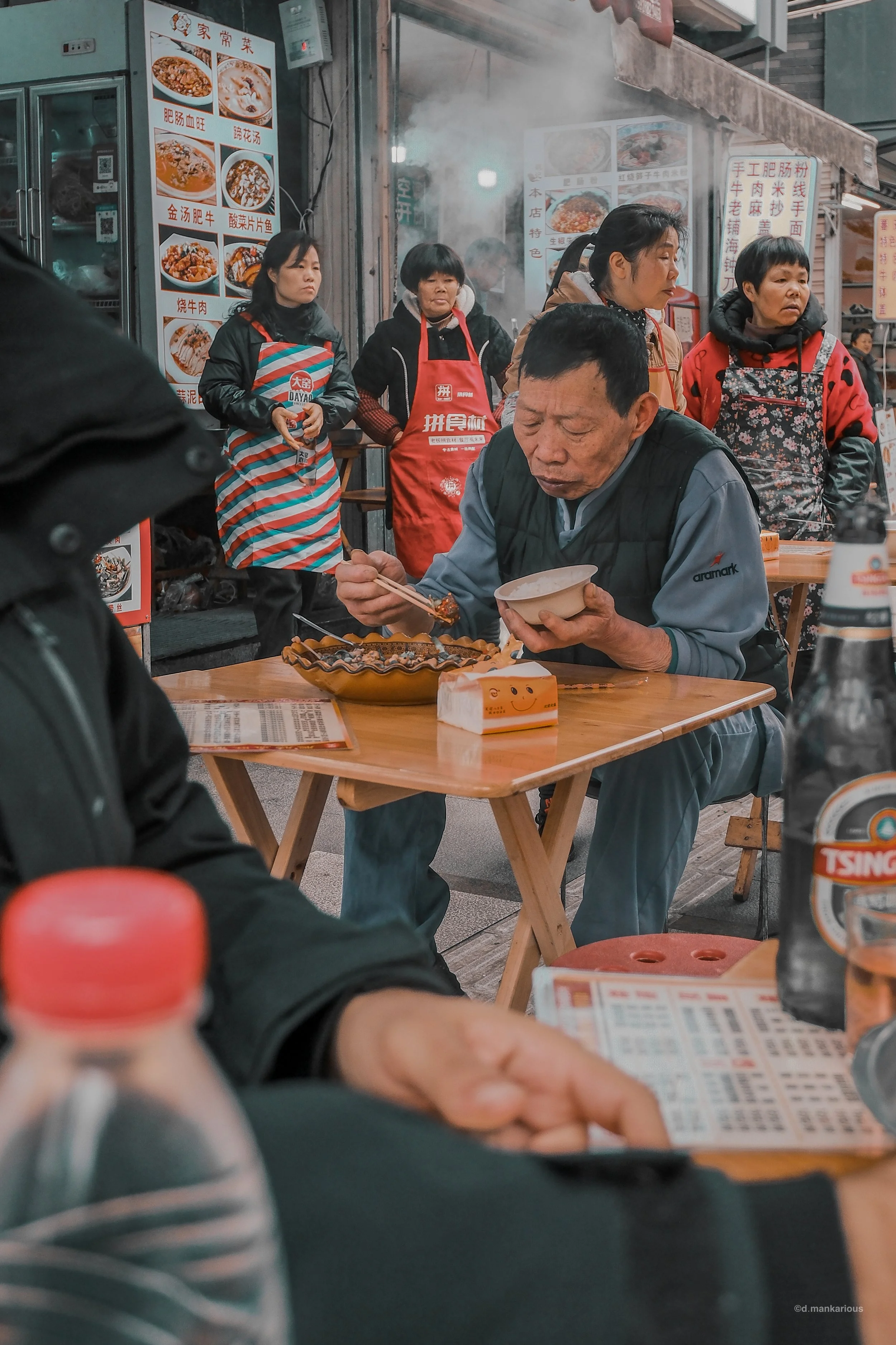An elderly man eating food with chopsticks at an outdoor restaurant, surrounded by other people and menu signs in Chinese.
