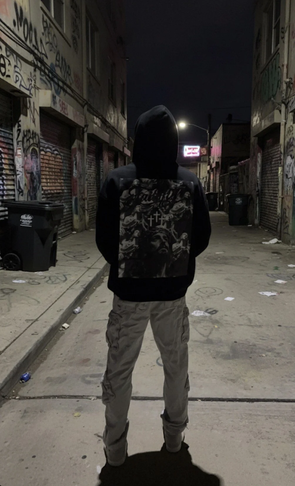 A person wearing a black hoodie and beige cargo pants standing in an urban alley at night, with graffiti-covered walls and closed storefronts on either side.