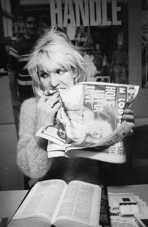 A woman with short, tousled blonde hair is sitting at a table, holding a folded newspaper near her face and looking to the side. An open book and some papers are on the table in front of her. In the background, there are a few people and a sign with the word "HANDLE" visible.