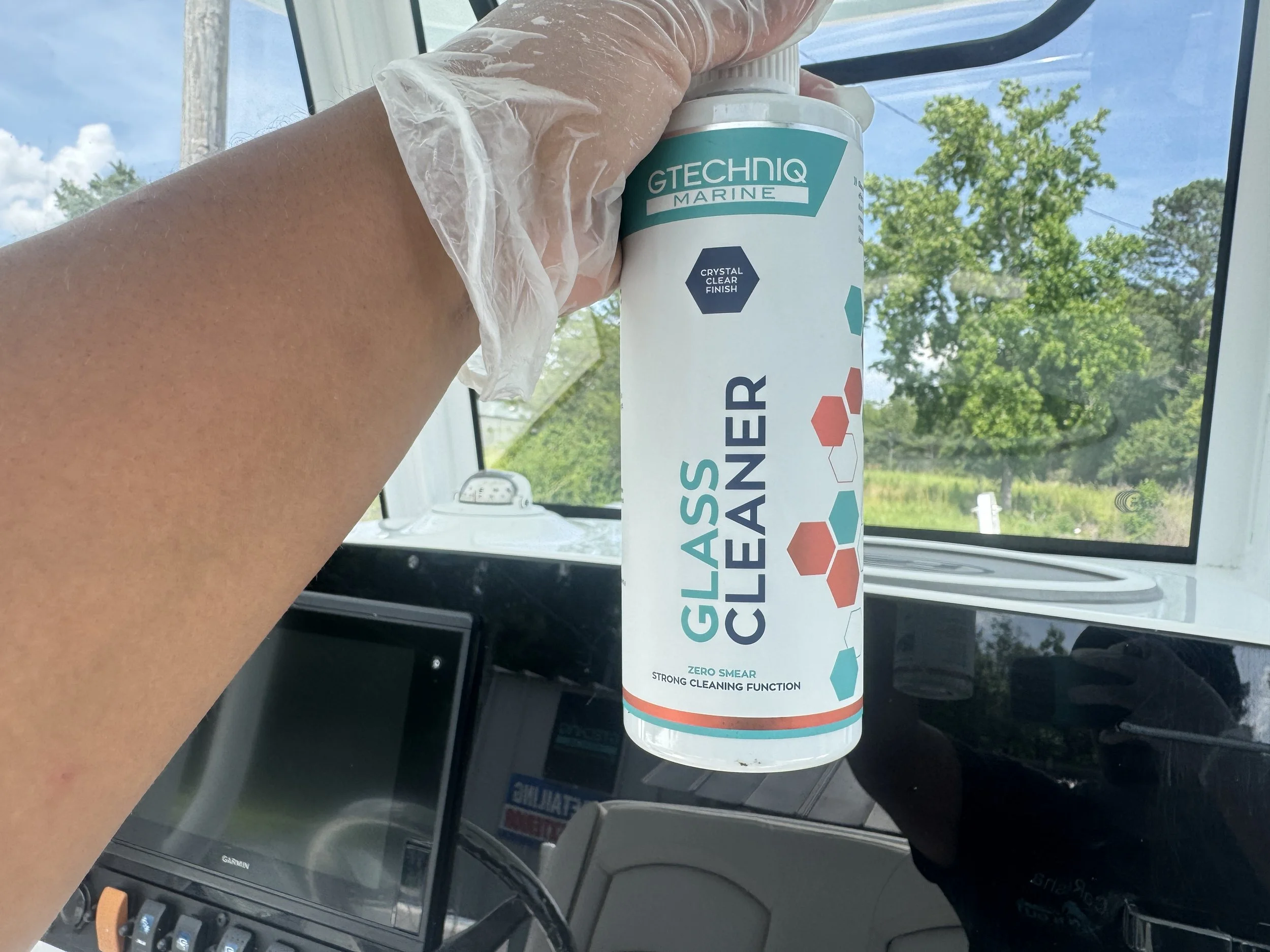 A person wearing a plastic glove holding a bottle of Gtechniq Marine Glass Cleaner inside a vehicle with a dashboard and a window showing green trees and blue sky outside.