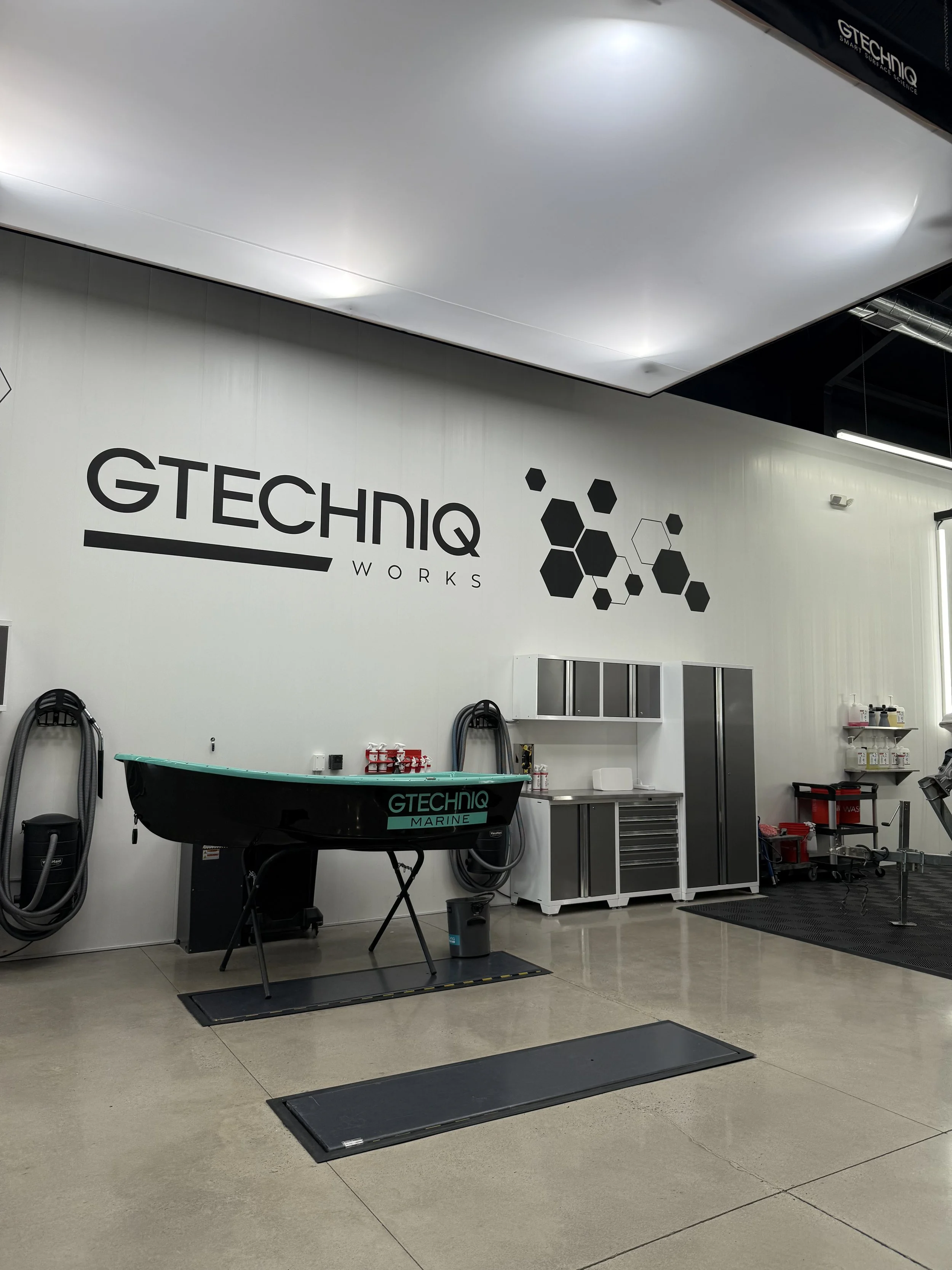 Industrial workspace with a boat on stands, storage cabinets, and wall decorations including a sign reading 'GTECHNQ WORKS' and hexagon patterns.