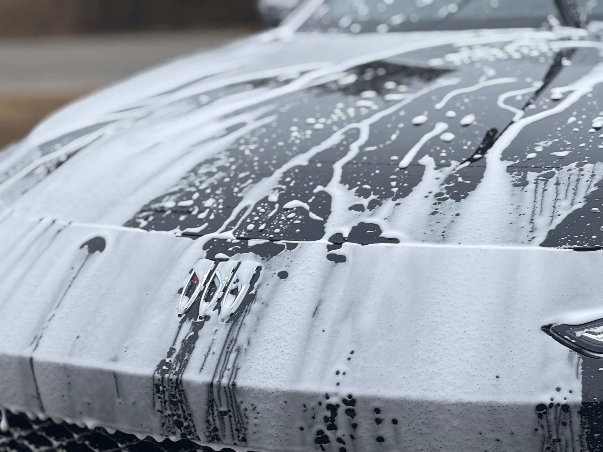 Close-up of a car hood covered in soap suds during a wash, with the hood emblem visible, and soap drips on the surface. Gtechniq decon wash 