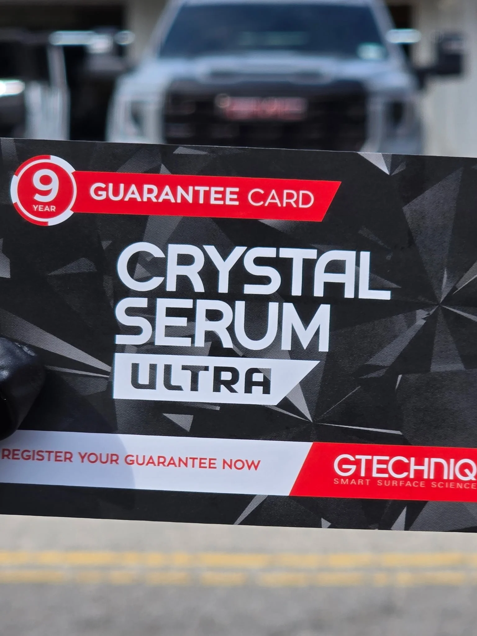 Close-up of a black and red certification card for Gtechniq's Crystal Serum Ultra, with part of a vehicle and street in the background.