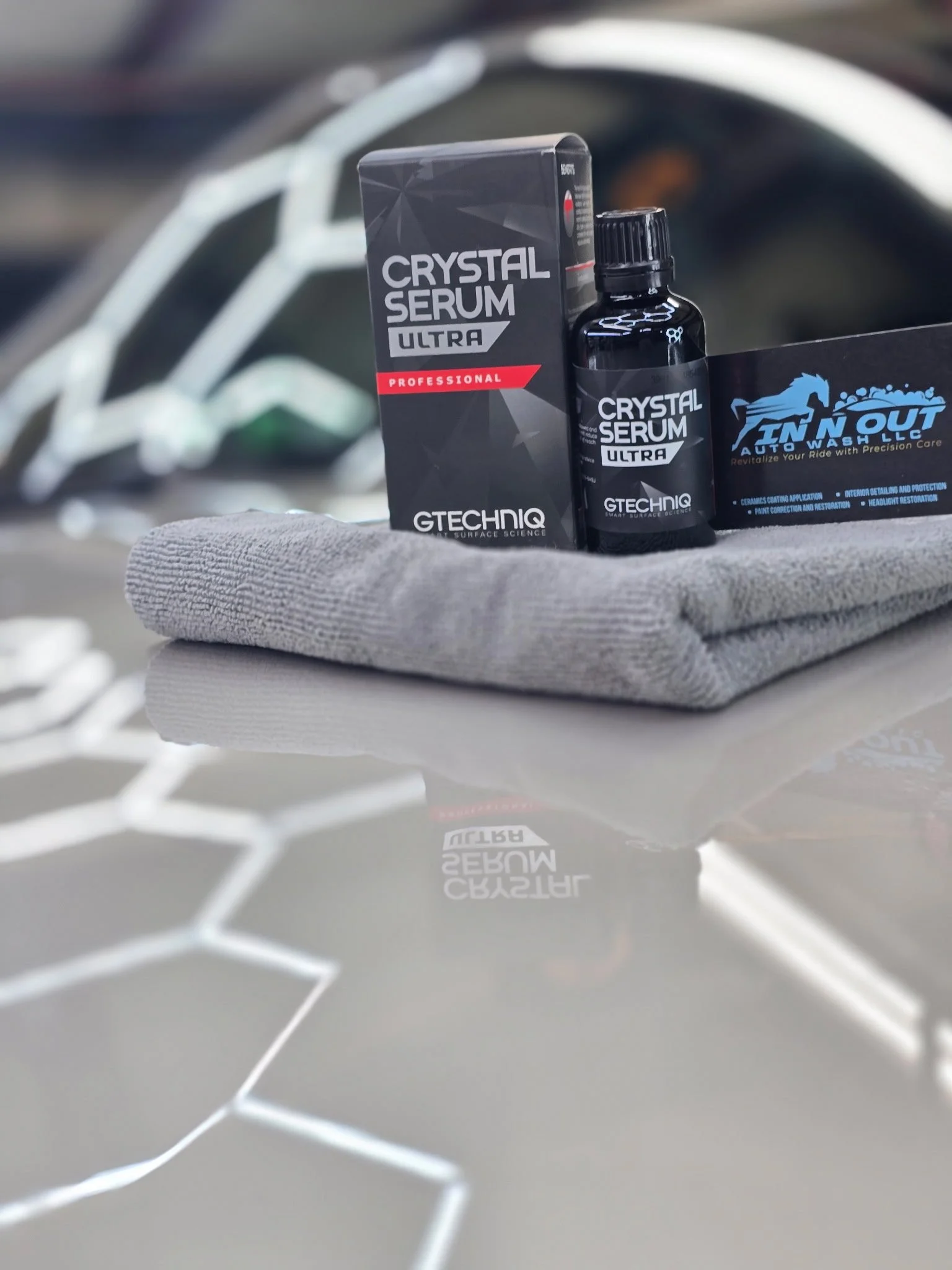 A bottle of GTECHNIQ Crystal Serum Ultra, its box, and a cloth are placed on a car surface for detailing, with a blurred background of a car wheel.
