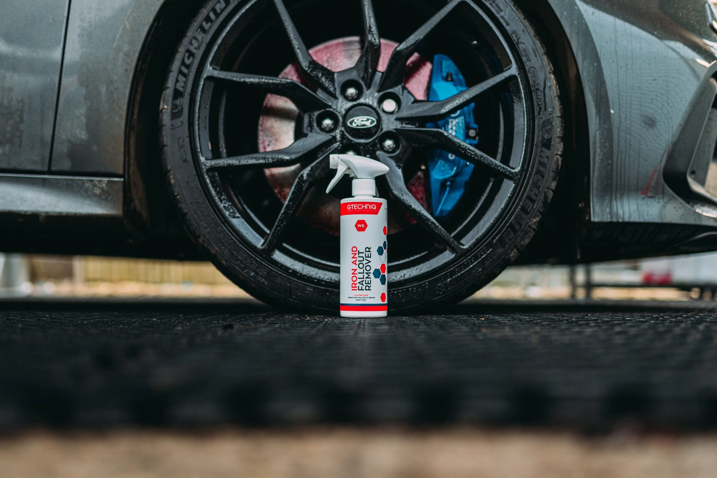 A spray bottle labeled 'Iron and Fall Out Remover' placed on the ground in front of a black car tire.