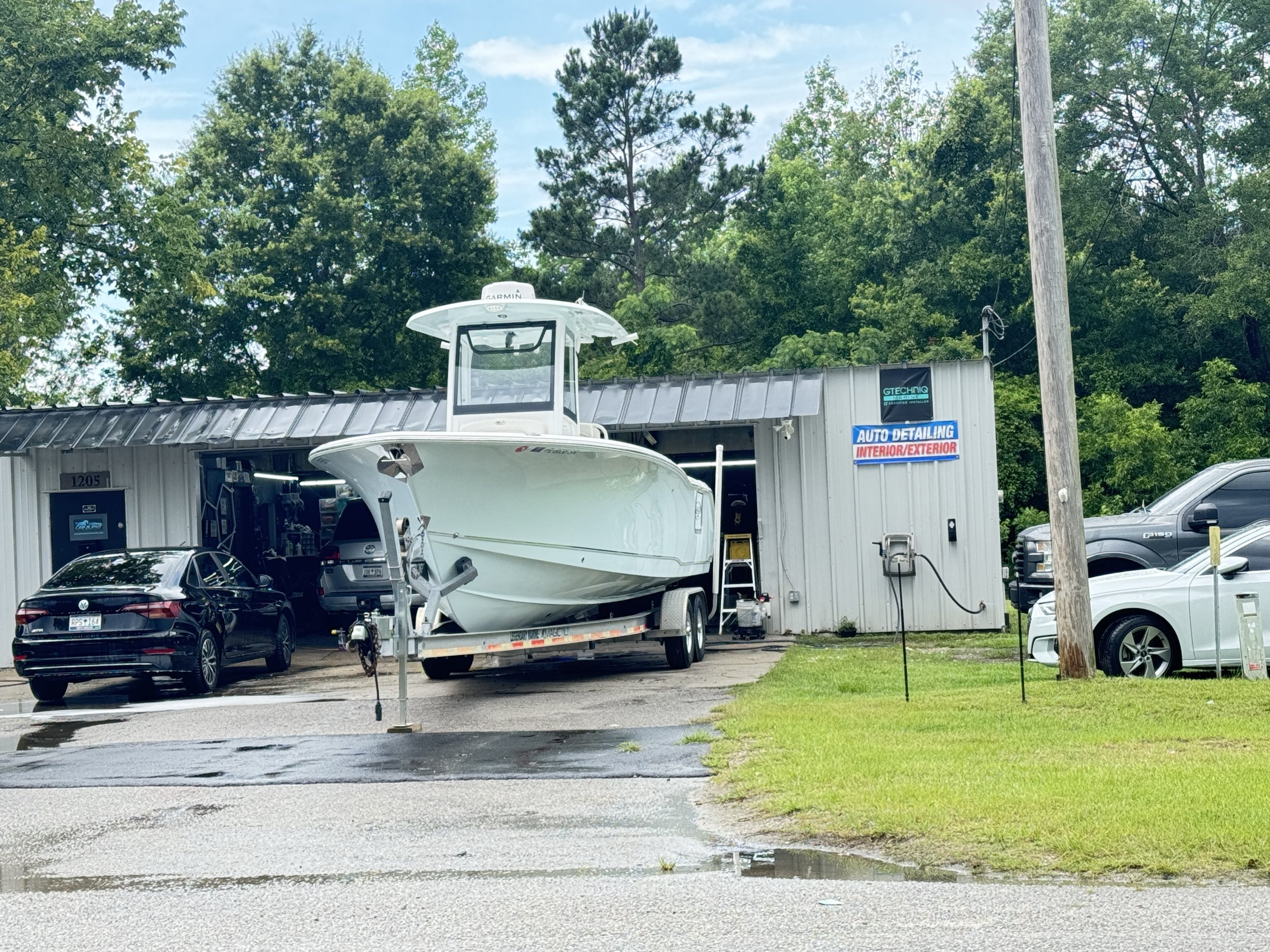 Auto detailing shop with a boat on a trailer, parked next to cars, with a building in the background and trees behind.