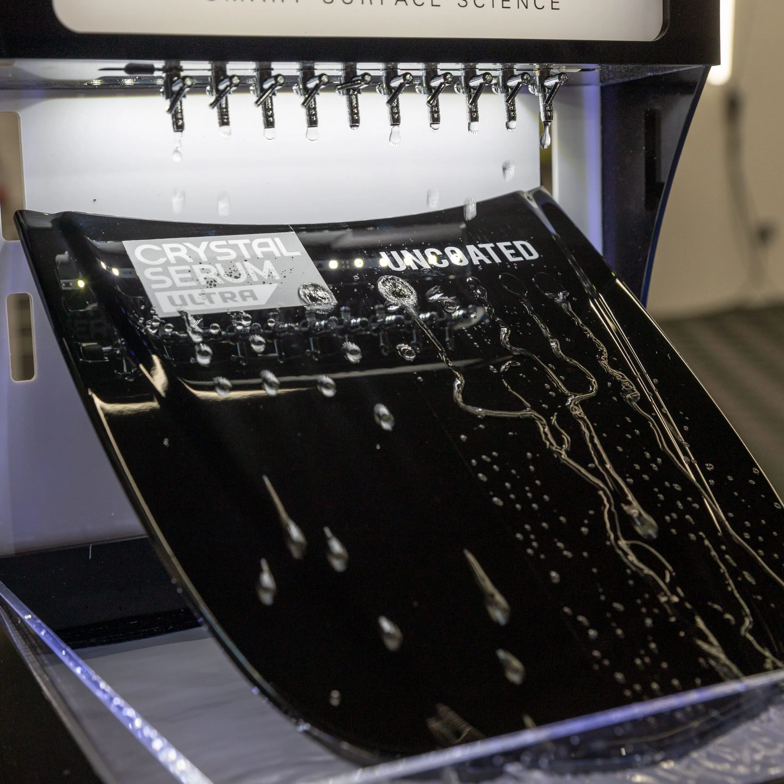 A black sheet of crystal serum ultra skincare product being processed in an automatic machine with multiple nozzles spraying liquid onto it. The sheet has labels 'CRYSTAL SERUM ULTRA' and 'UNC8ATED' visible on it.