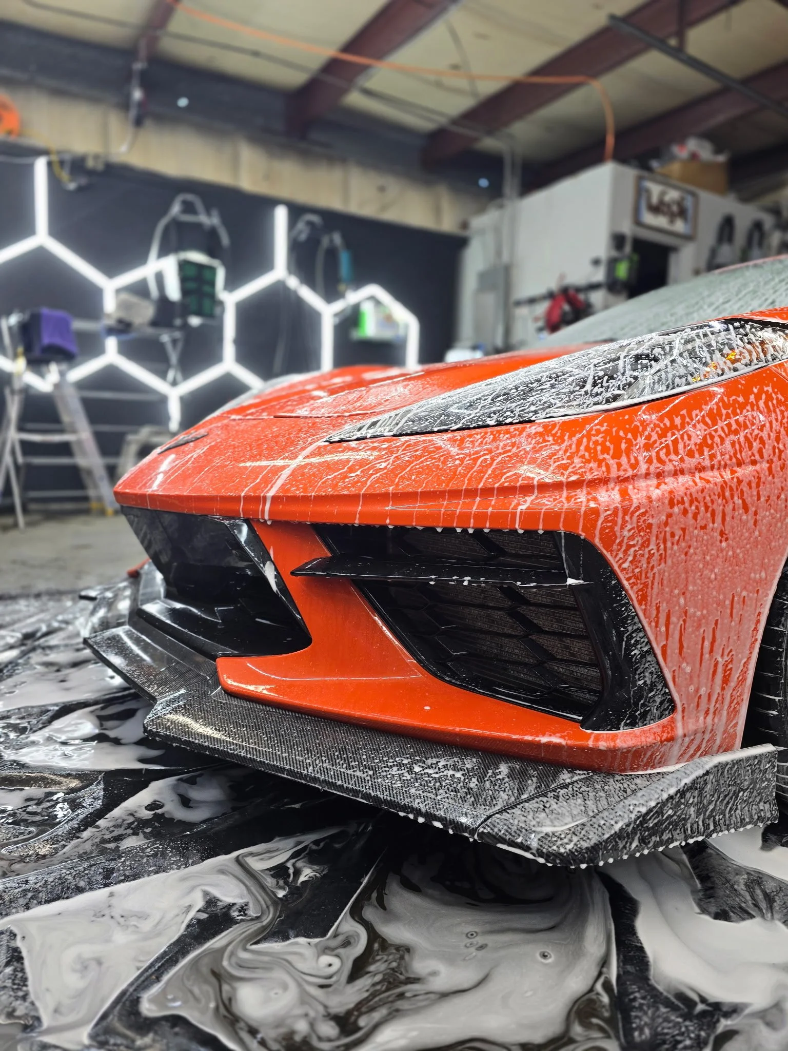 Close-up of an orange sports car with soap and water on its surface, inside an automotive detailing or wash station.