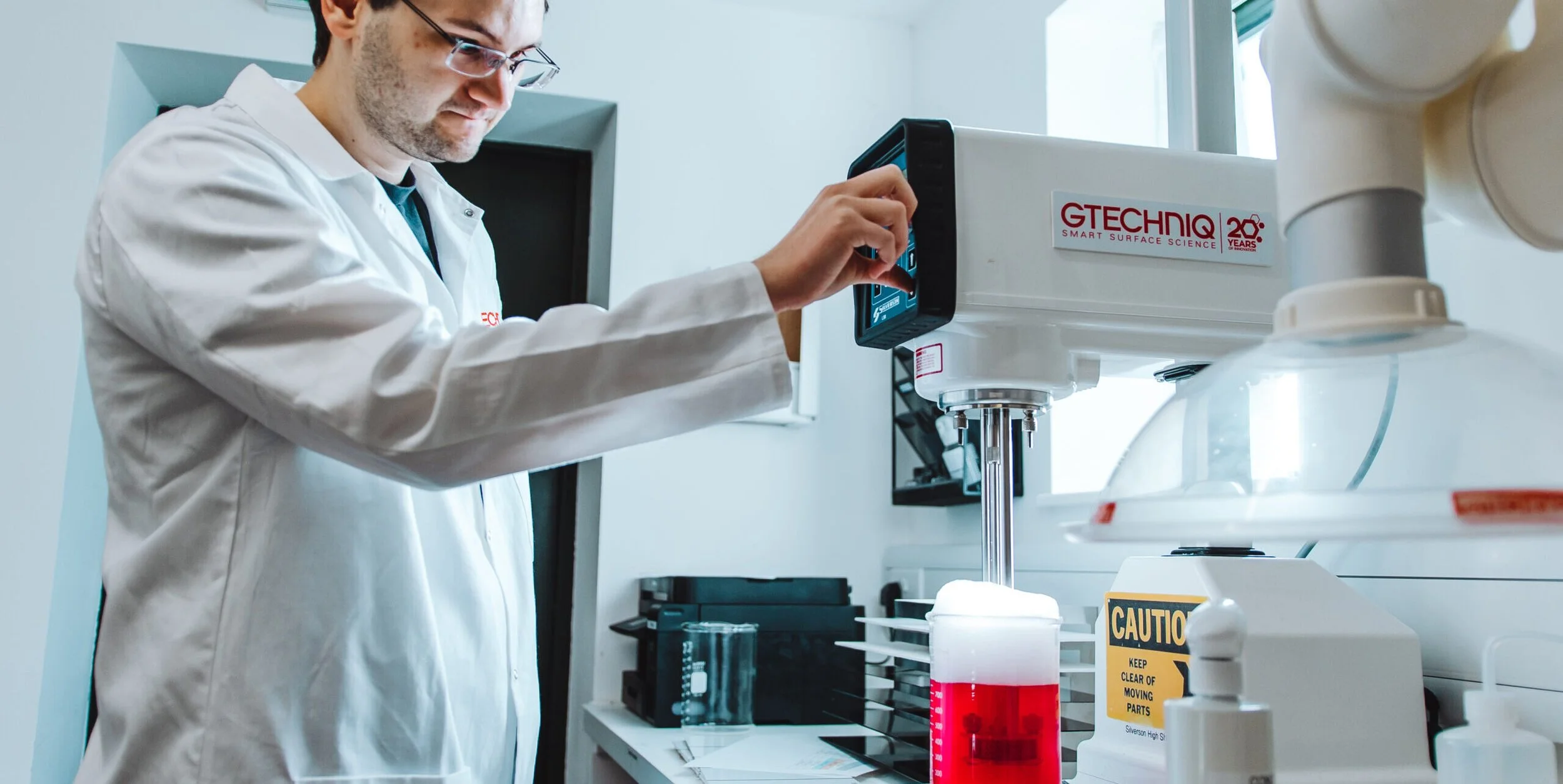 A scientist in a lab coat adjusts a machine in a laboratory with test tubes, equipment, and caution signs in the background.