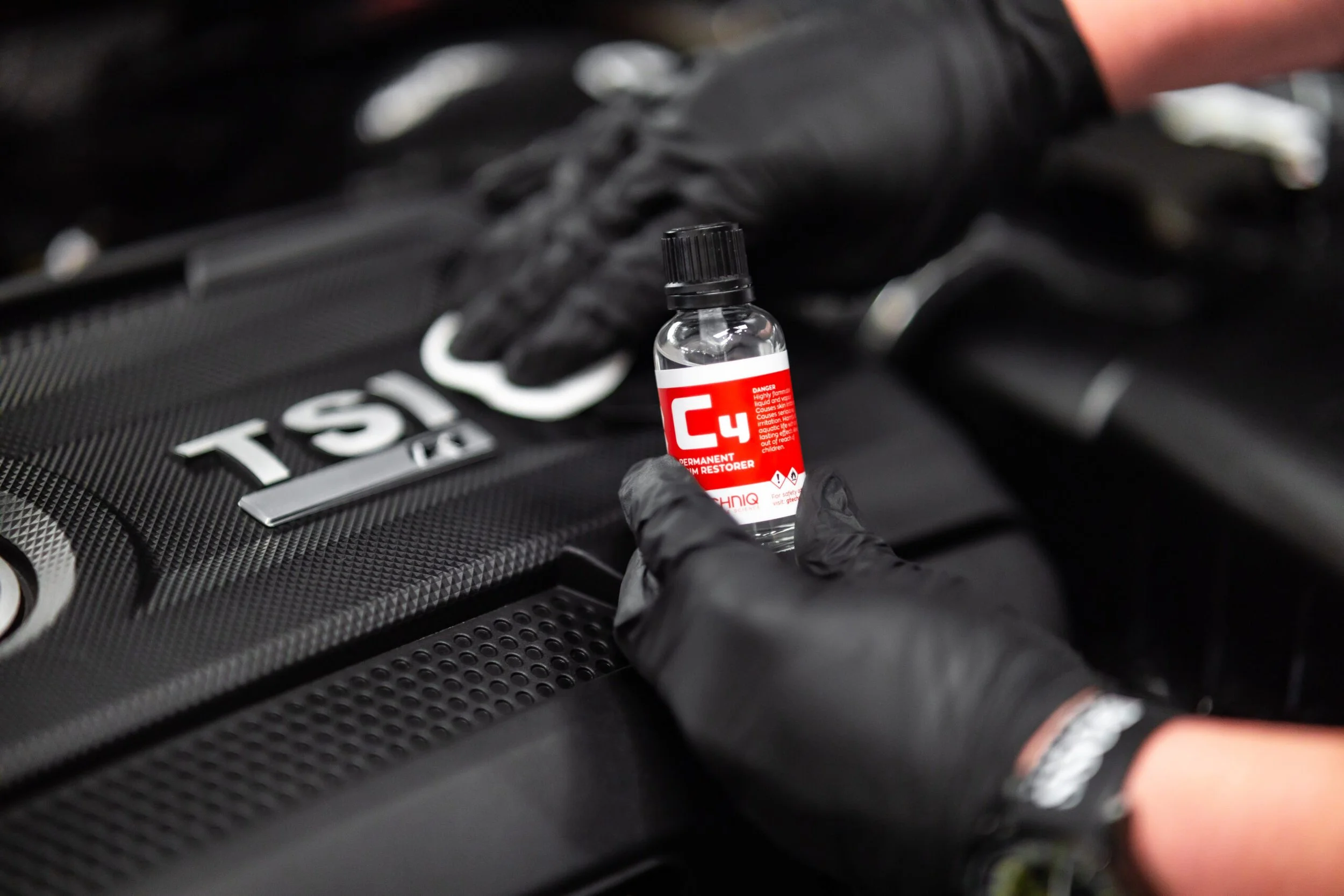 A person wearing black gloves is holding a small bottle labeled C4, a chemical used as a fuel restorer, near the engine of a car that has the TSI badge.