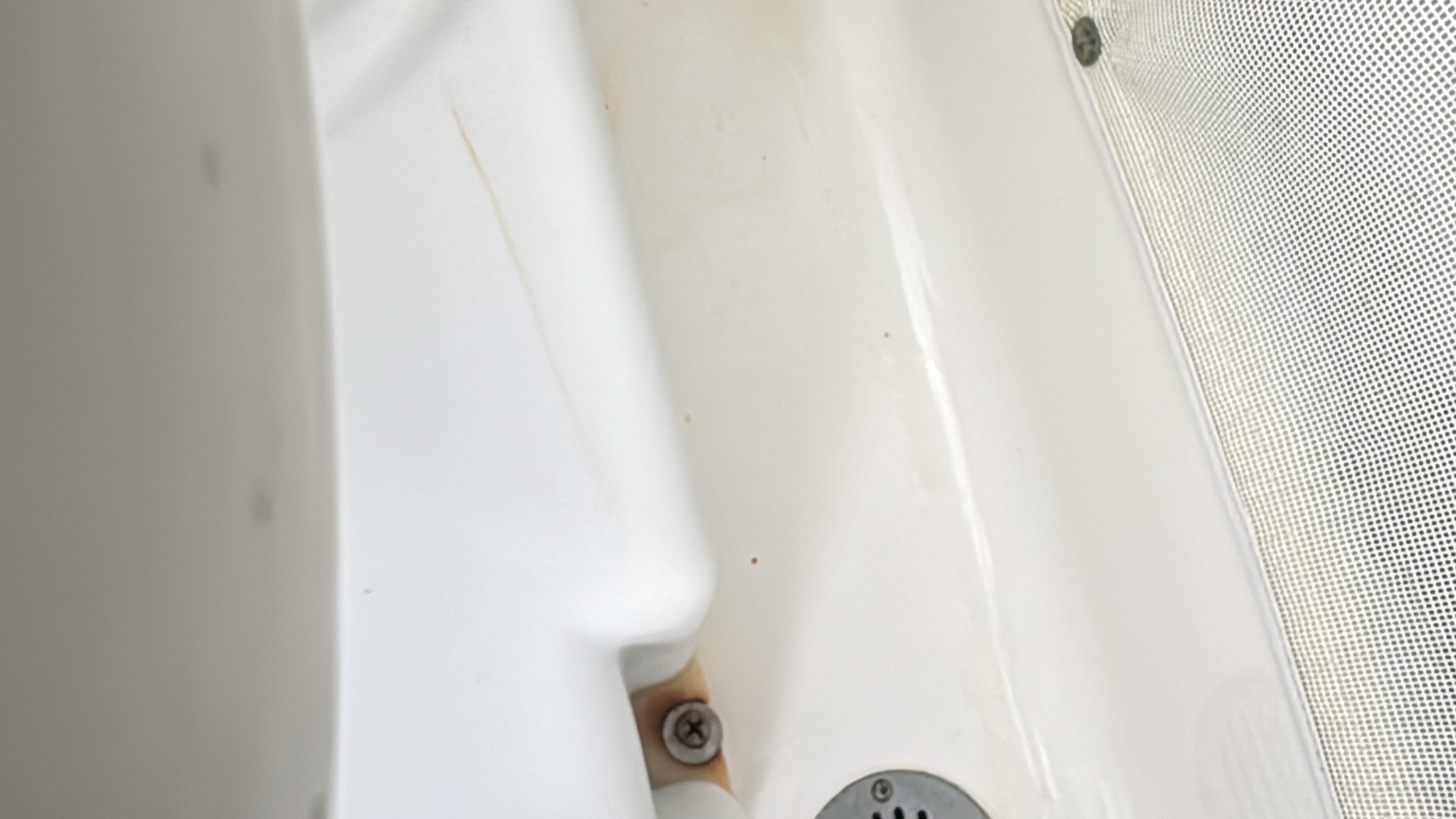 Close-up of a white bathtub with a metal drain and a visible screw, with some minor stains and a textured wall or screen nearby.