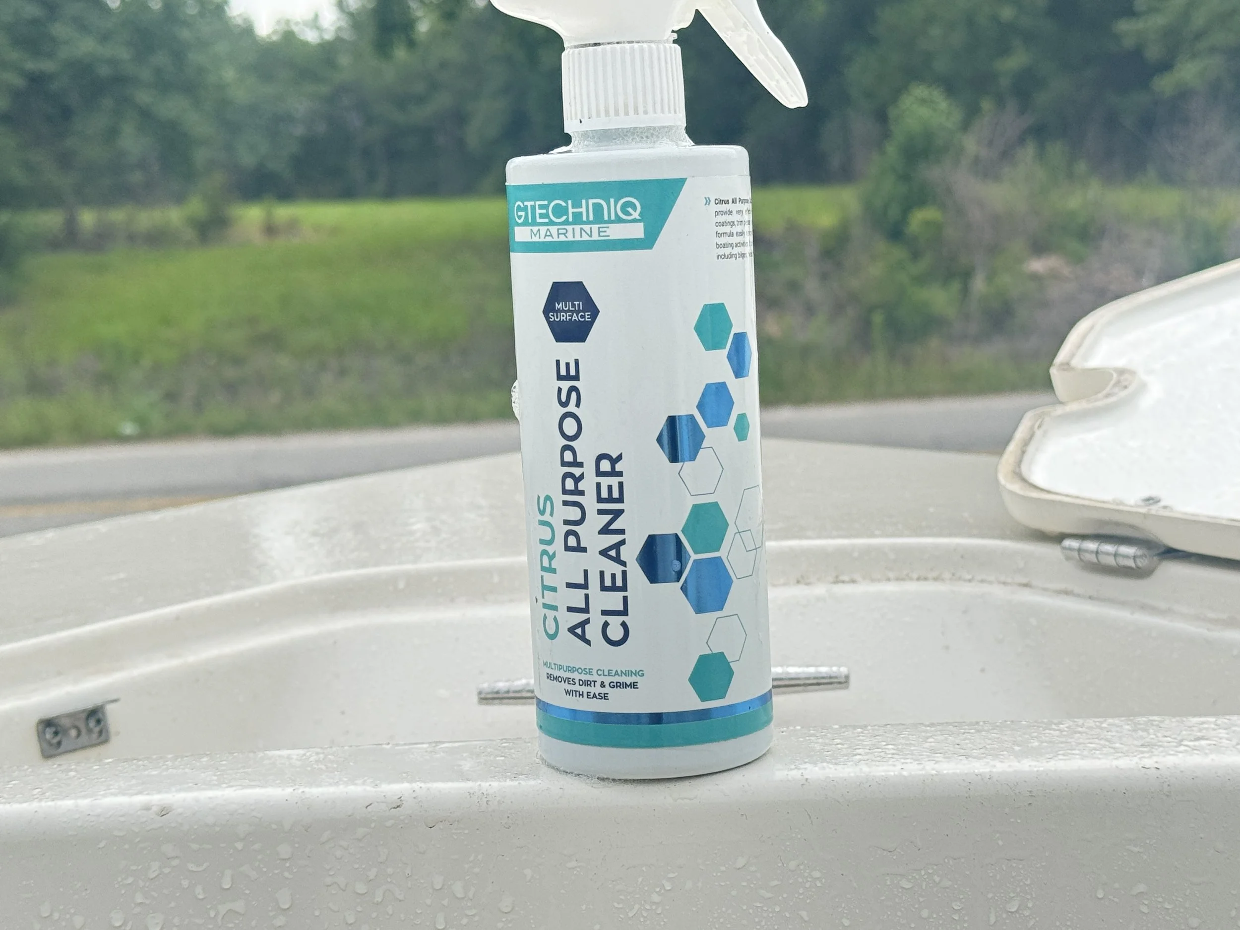 A bottle of multi-surface all purpose cleaner placed on a white boat or sink with water droplets, with a blurred outdoor greenery background.