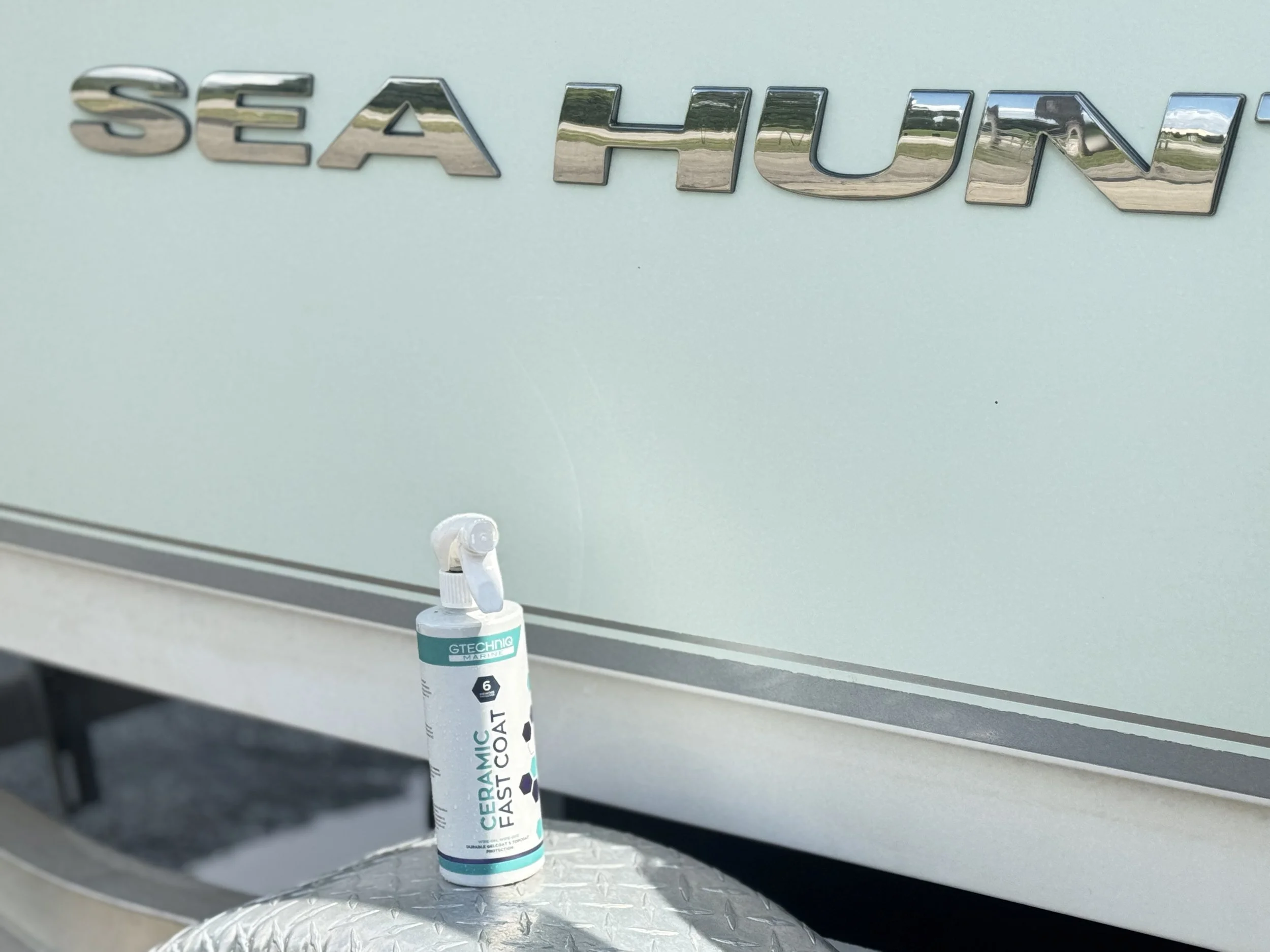 A bottle of ceramic fast coat spray resting on a metallic surface in front of an ice blue vehicle with a chrome 'Sea Hunt' emblem.