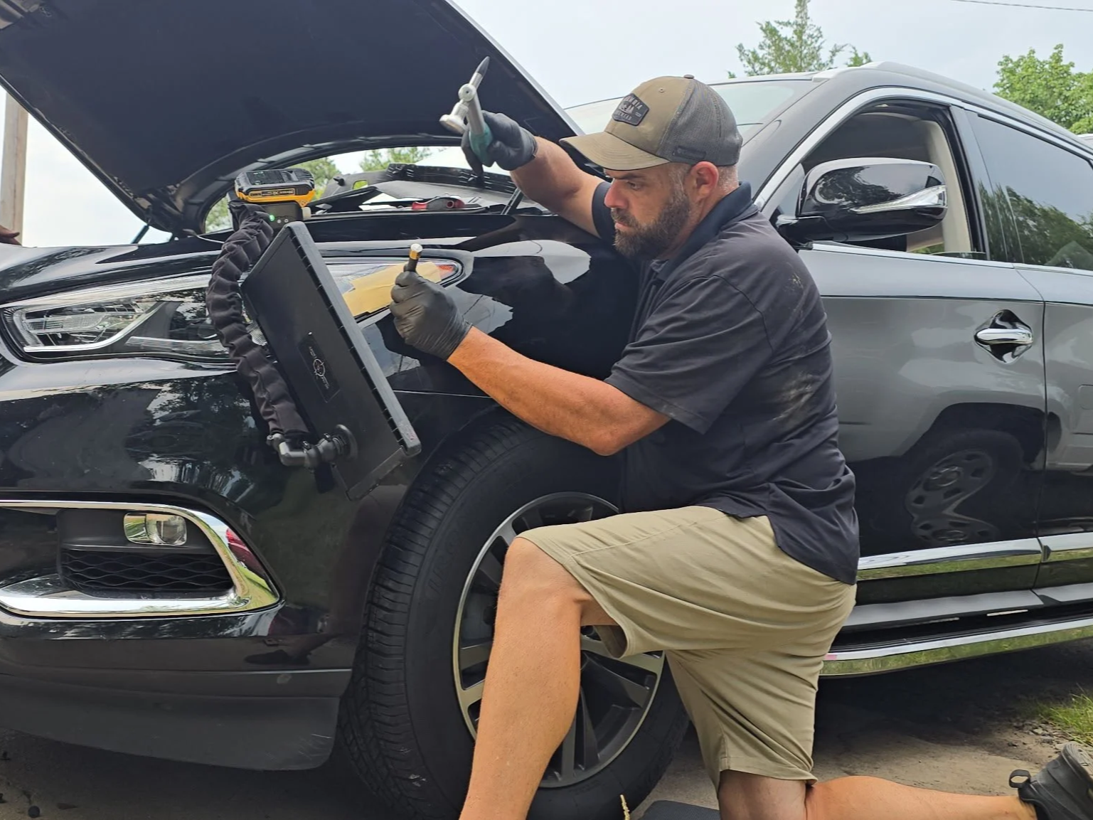 A man working on a black car with the hood open, using tools, with a clipboard or tablet on a stand, outside during daytime.