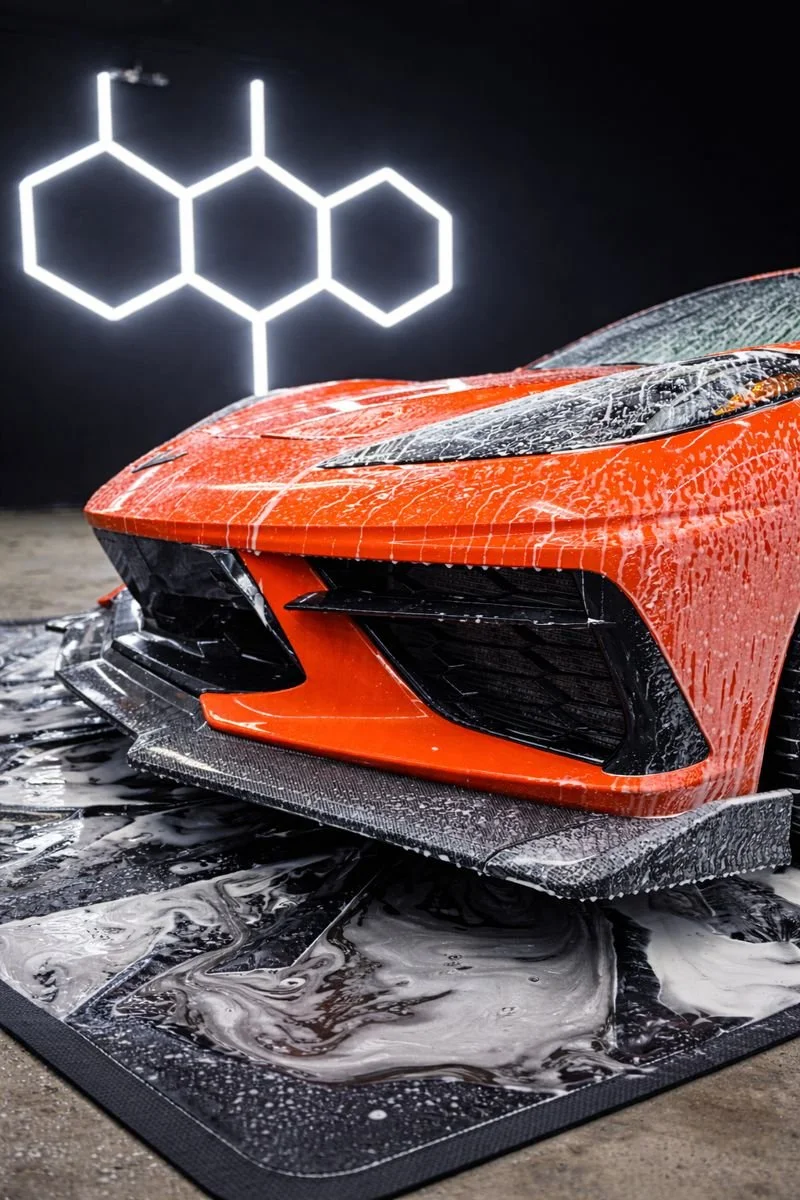 Close-up of a red sports car with a wet surface, on a cleaning mat, under a black wall with a hexagonal molecule design.