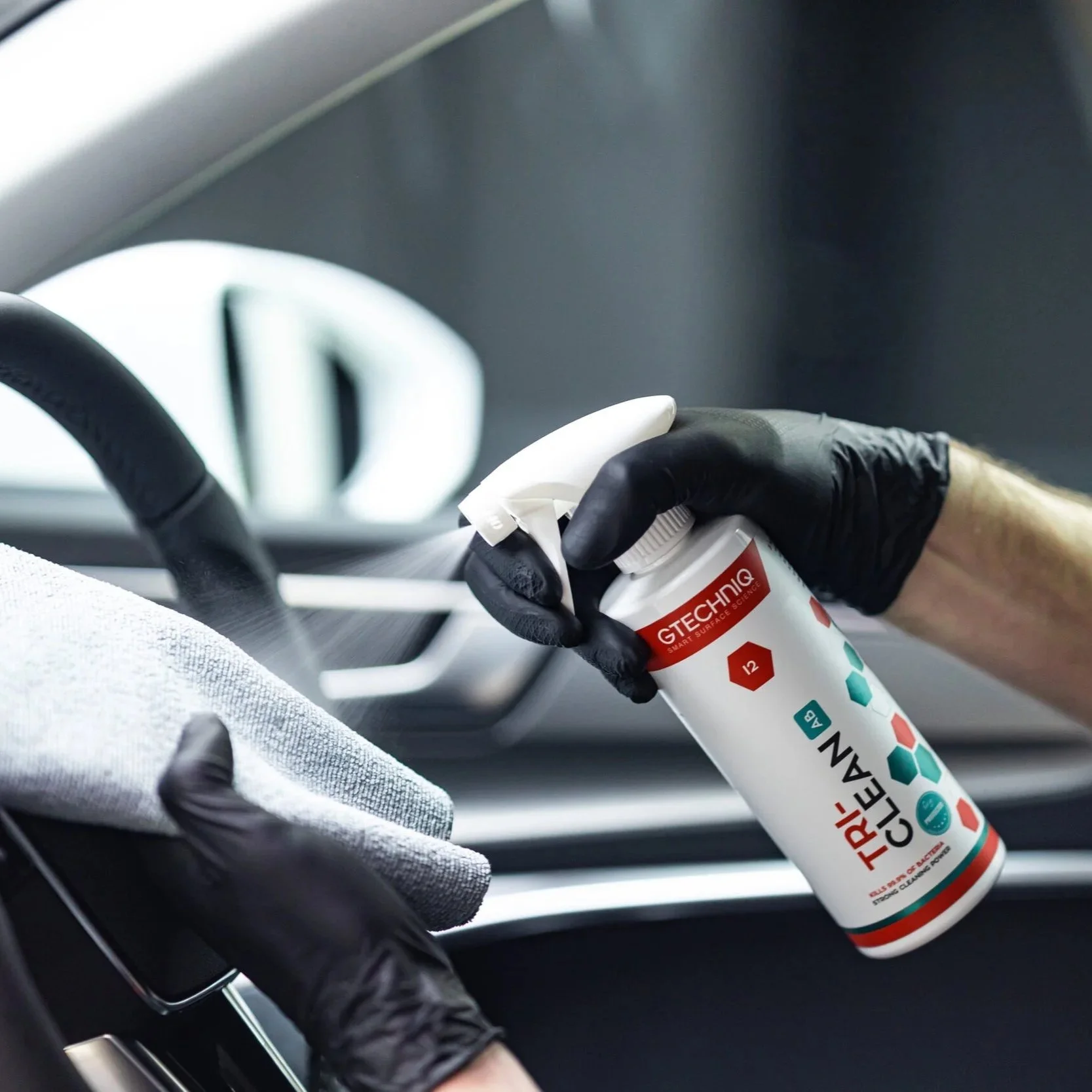 Person wearing black gloves using a spray bottle labeled 'TRI CLEAN' to disinfect a car interior surface.