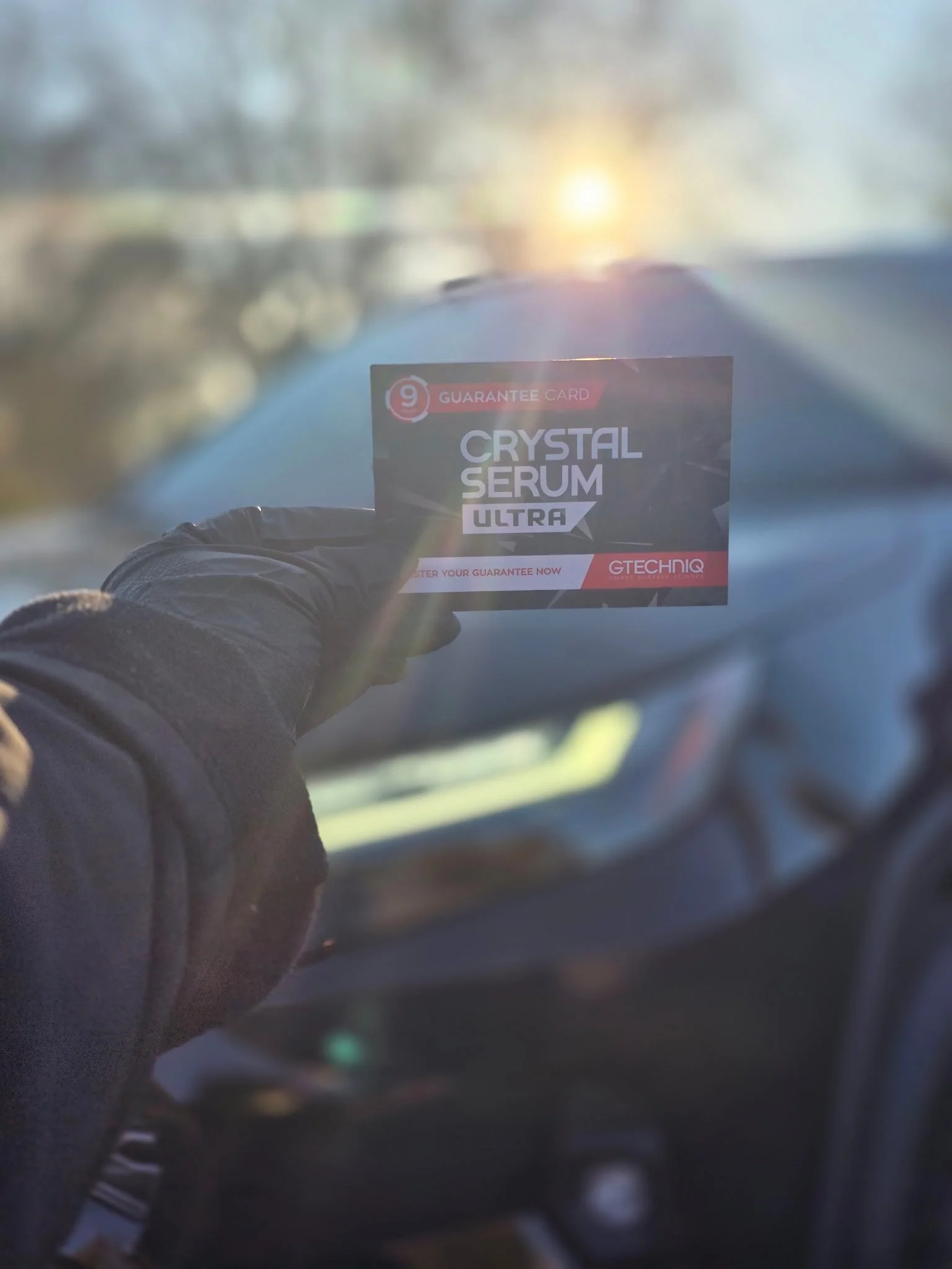 Person wearing black gloves holding a card labeled 'Crystal Serum Ultra' inside a vehicle with a blurred view of the outside at sunset.