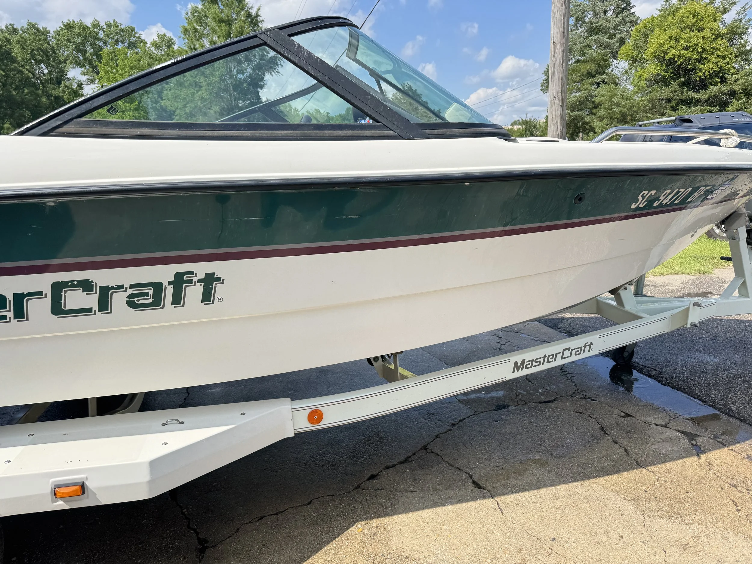 A green and white MasterCraft speedboat on a trailer parked on cracked asphalt, with trees and a utility pole in the background.