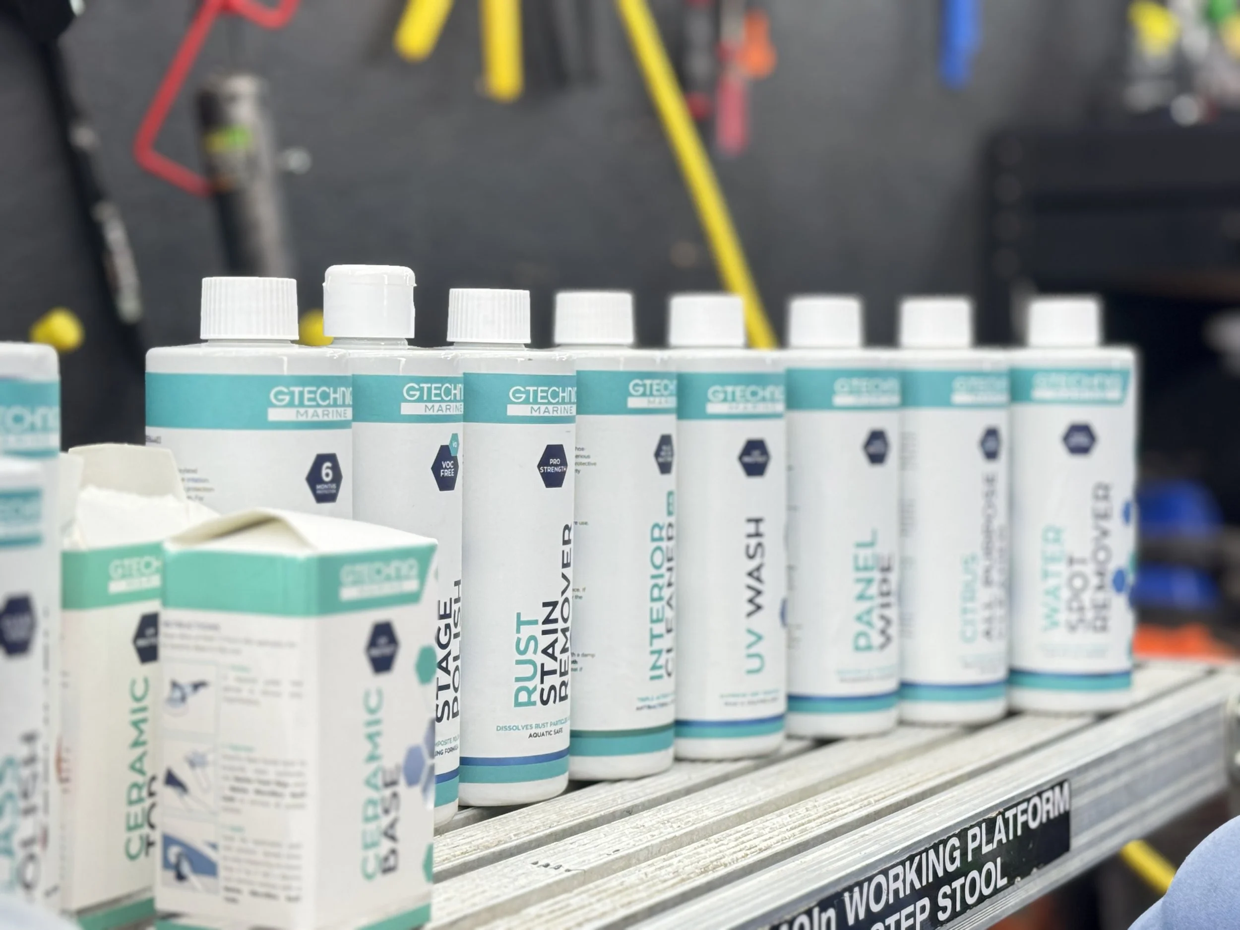 A row of white bottles with blue labels on a metal shelf, labeled with various cleaning and maintenance products, in a workshop setting.