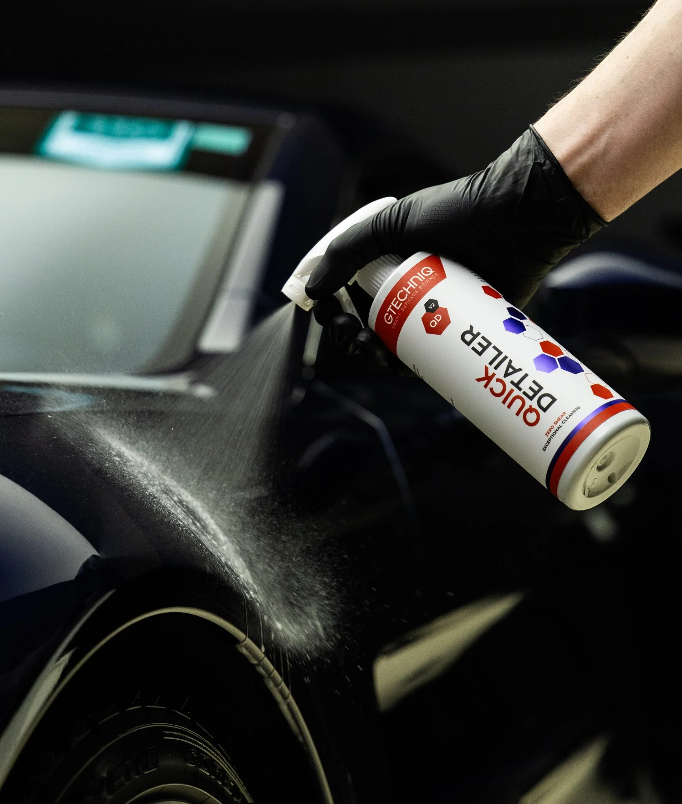 A person wearing a black glove using a can of quick detailer spray on a car's surface, with a spray pattern visible on the vehicle's black painted surface.
