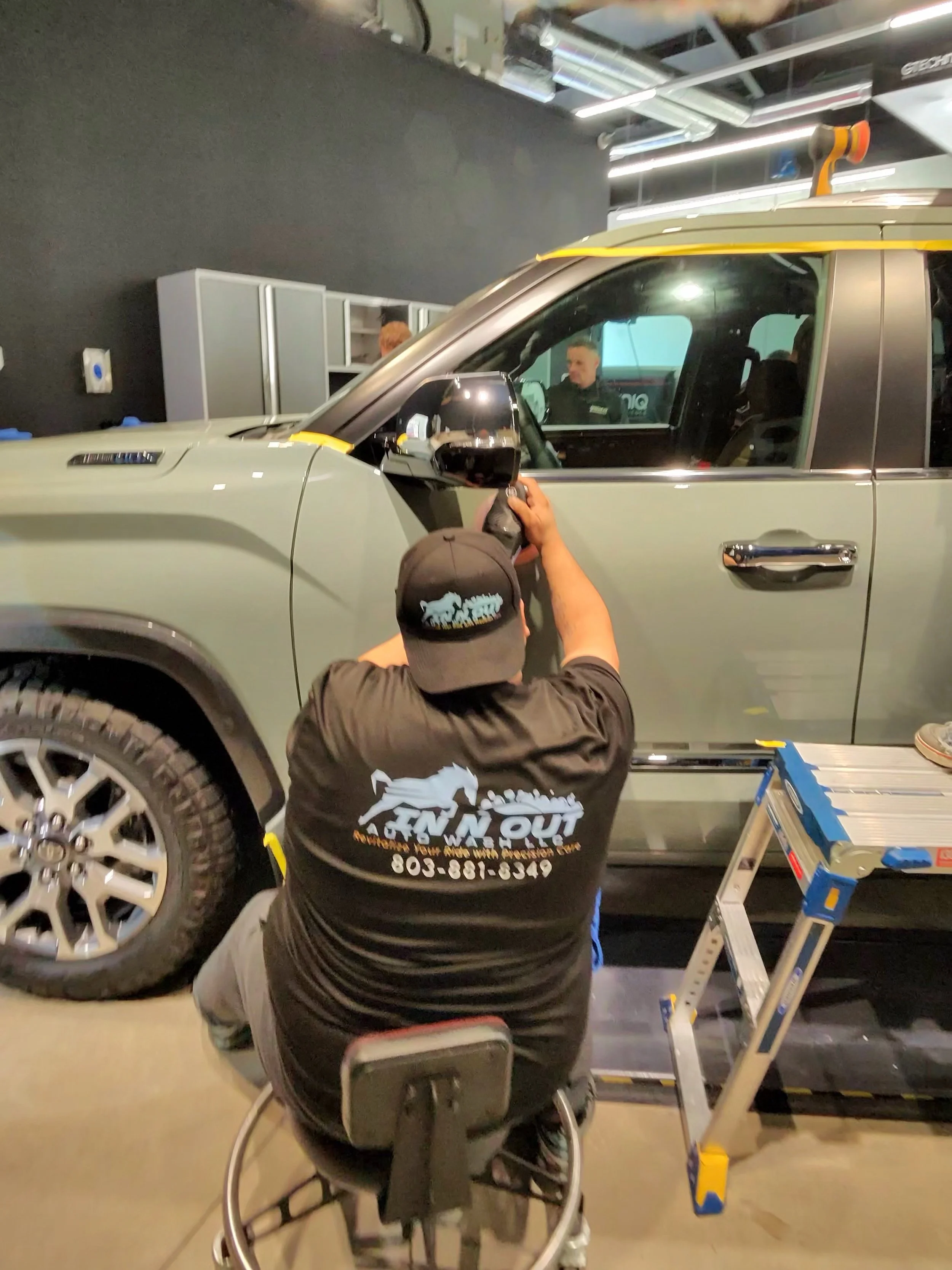 Technician in a black shirt and cap with the logo 'FNN OUT' cleaning or inspecting a grey pickup truck at an auto repair shop or detailing garage.