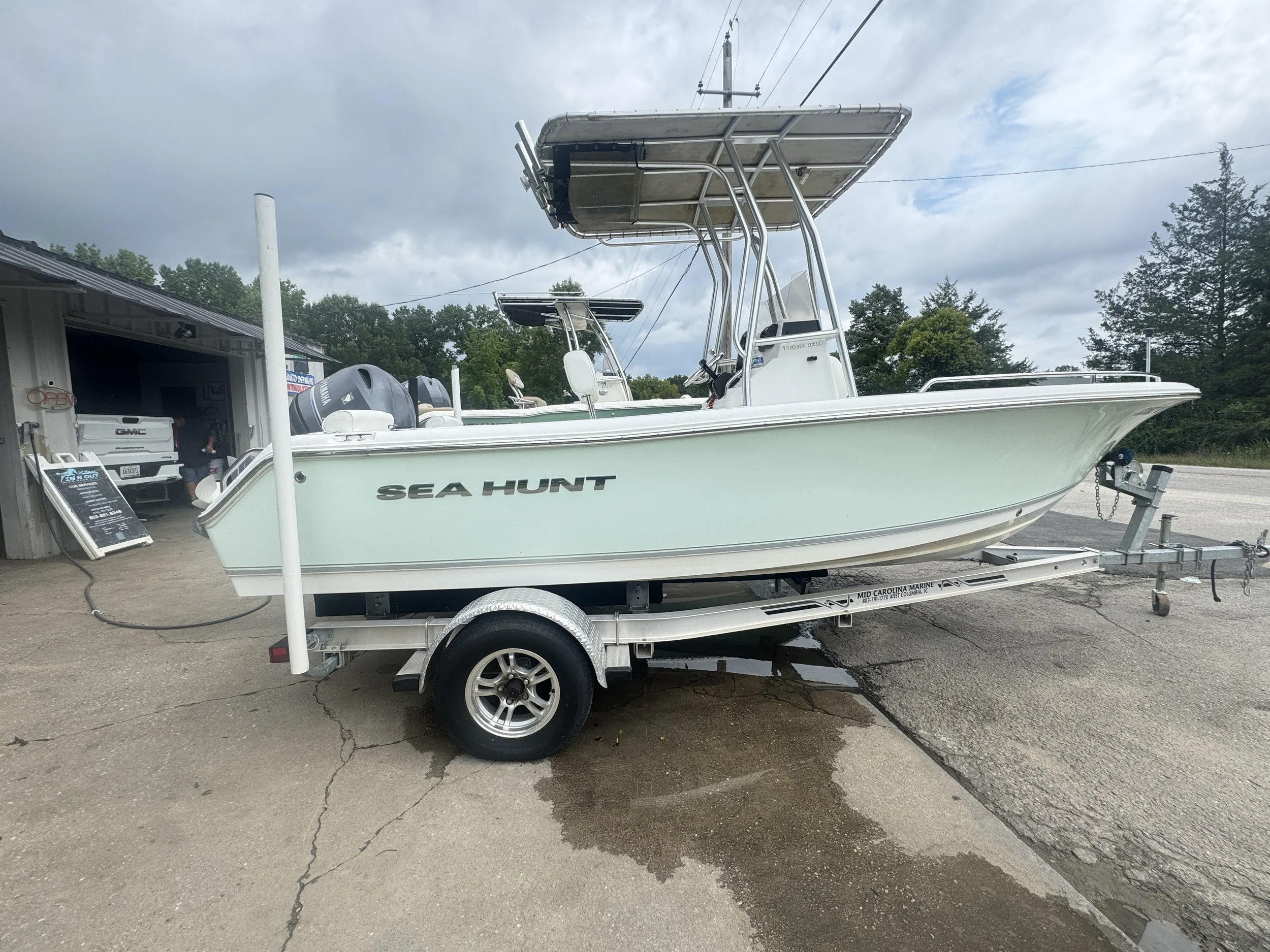 A light blue Sea Hunt boat on a trailer near a building with a boat shop, with trees and a cloudy sky in the background.
