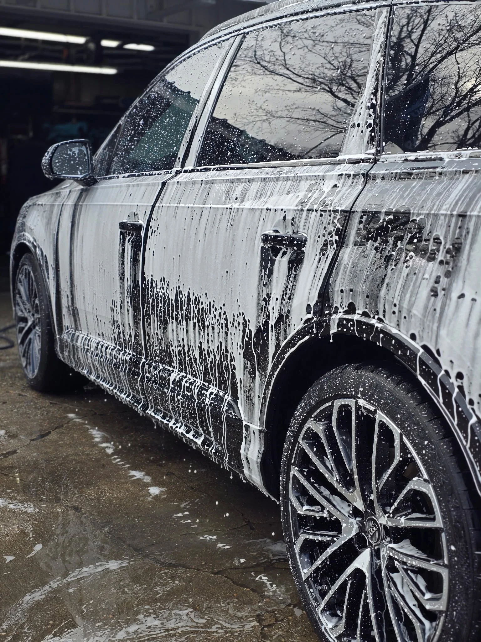 A black car being washed with soap, with foam covering the sides and wheels, outdoors on a concrete surface.