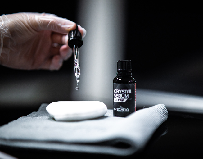 A hand holding a dropper releasing a drop of serum into a white dish, with a bottle labeled "Crystal Serum Ultra" nearby, placed on a gray cloth.