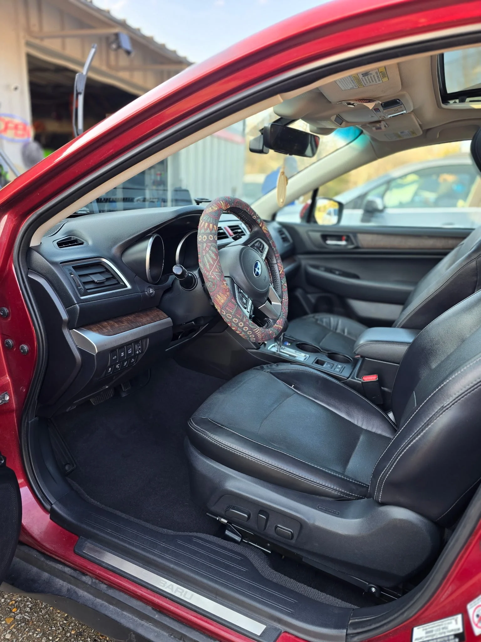 Red Subaru car interior showing black leather seats, a steering wheel with a patterned cover, dashboard, and center console, parked outdoors.